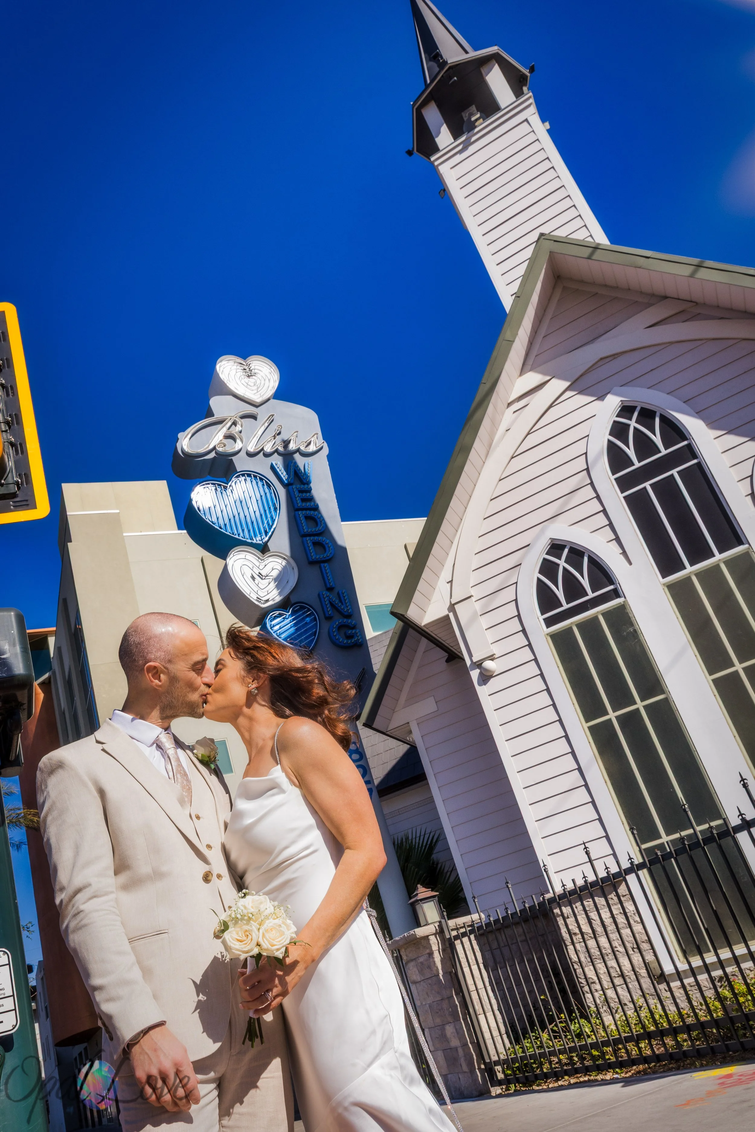 Bride and groom kissing outside Bliss Wedding Chapel under a bright blue Las Vegas sky.