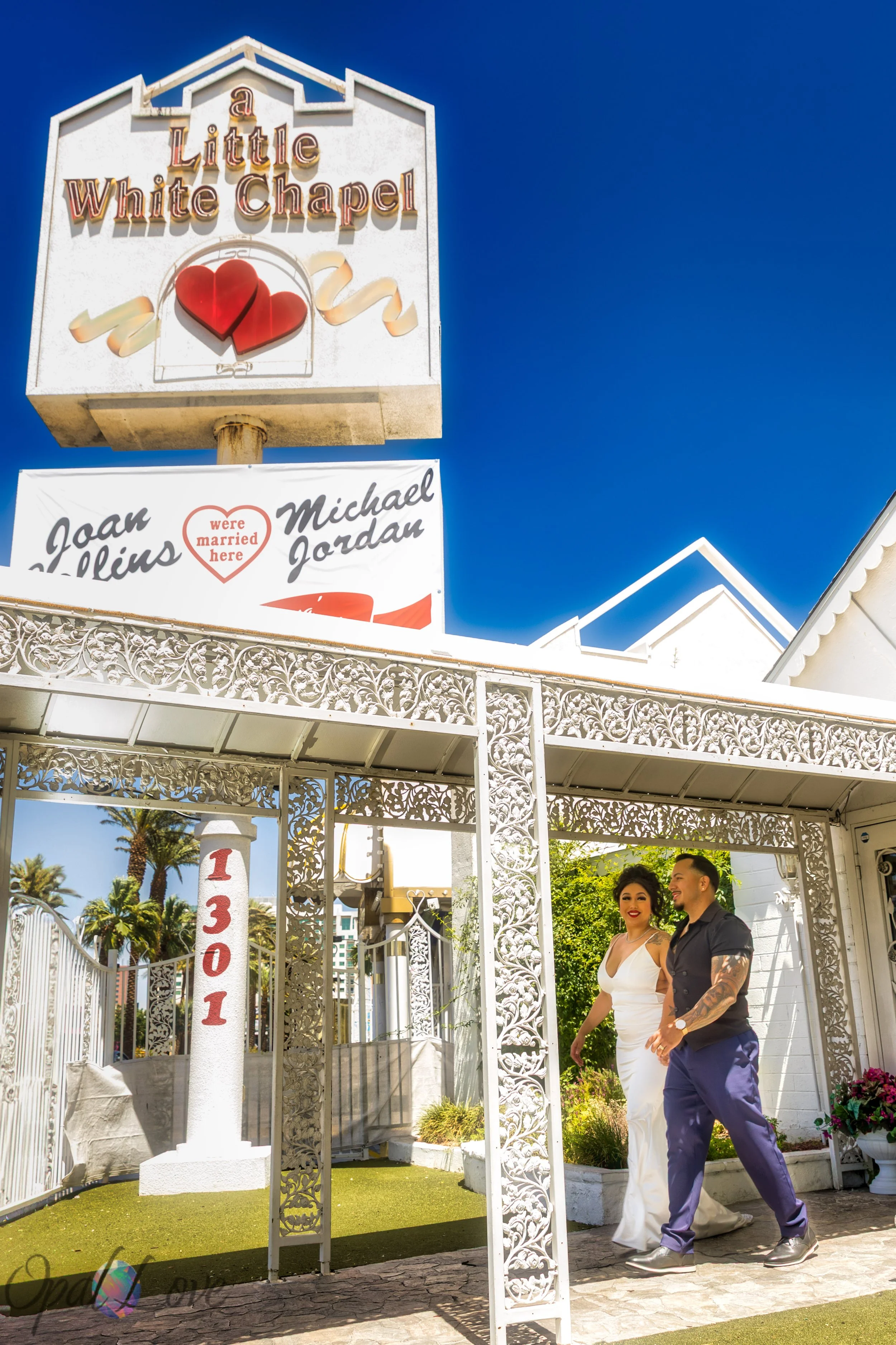 

Newlyweds walking hand in hand beneath the Little White Chapel sign and heart-shaped canopy.