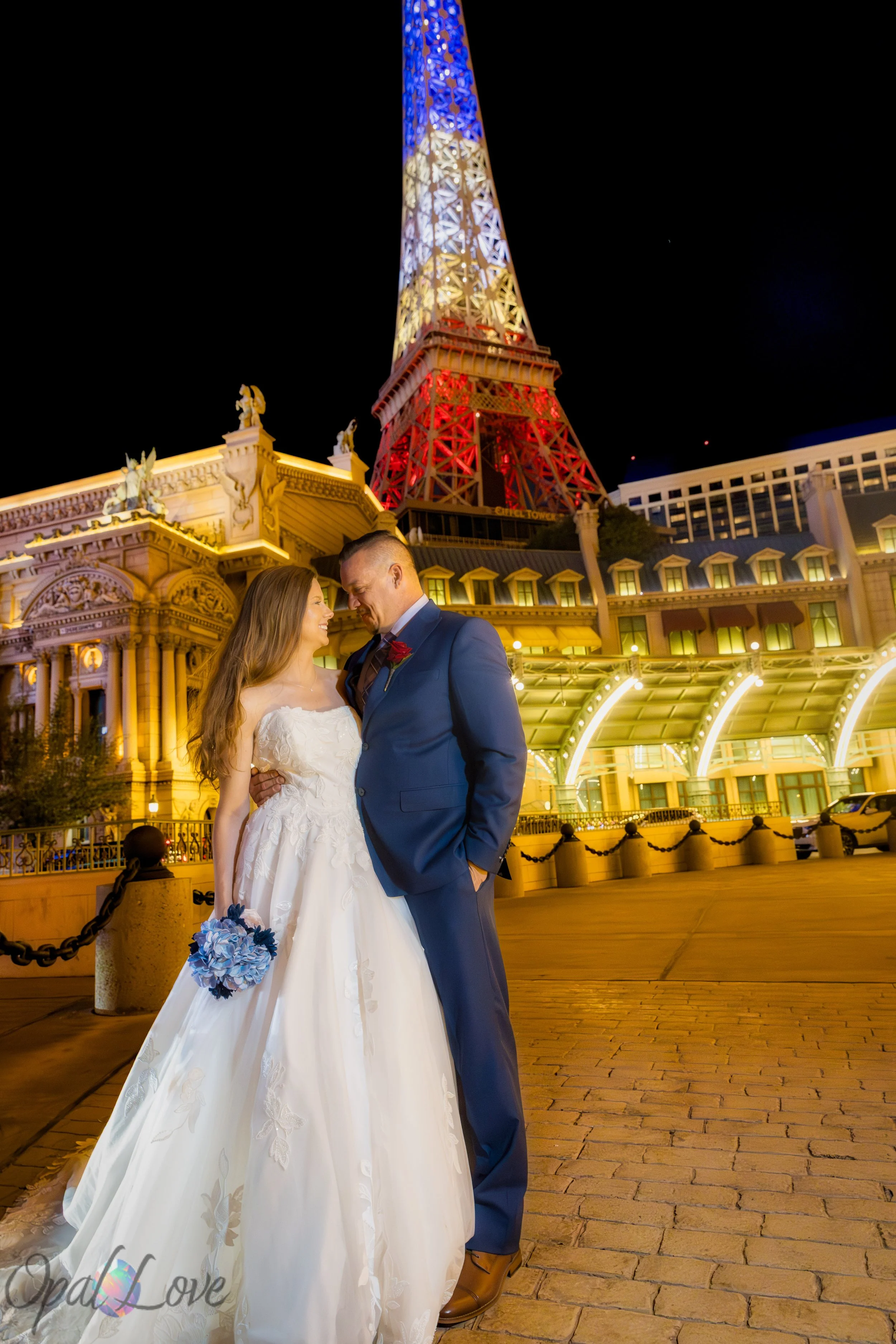 Couple standing together smiling with the illuminated Eiffel Tower at Paris Las Vegas in the background.