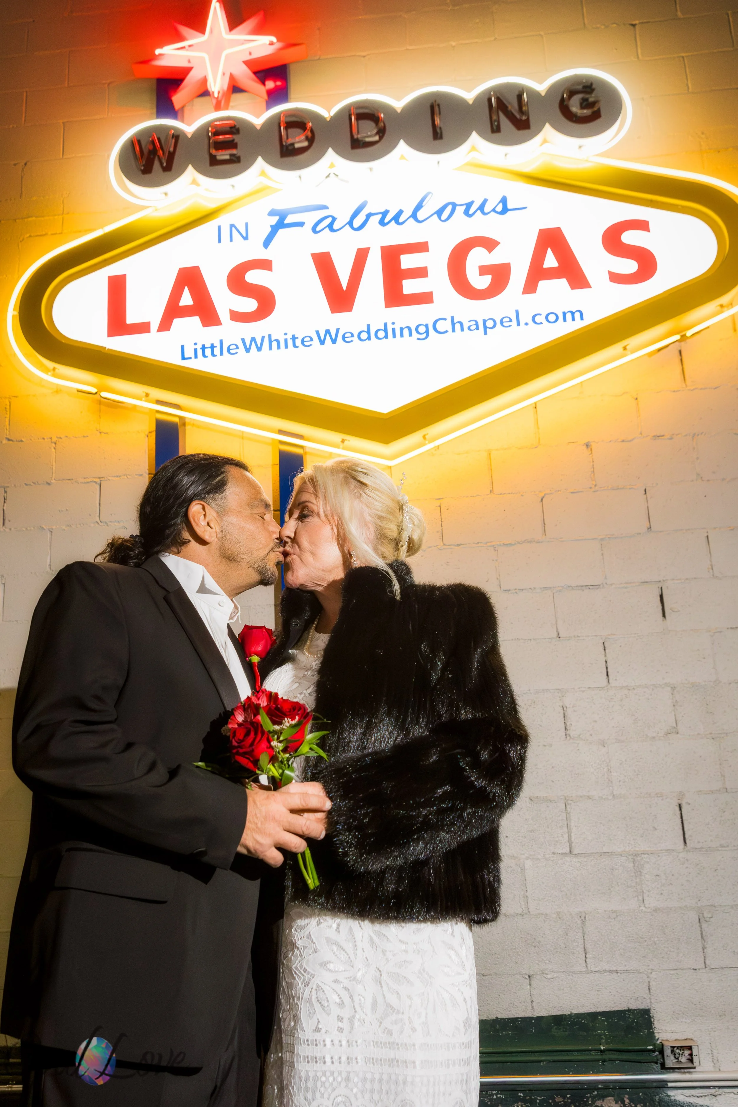 Bride and groom kissing under the Welcome to Las Vegas sign on the Little White Wedding Chapel grounds at night.