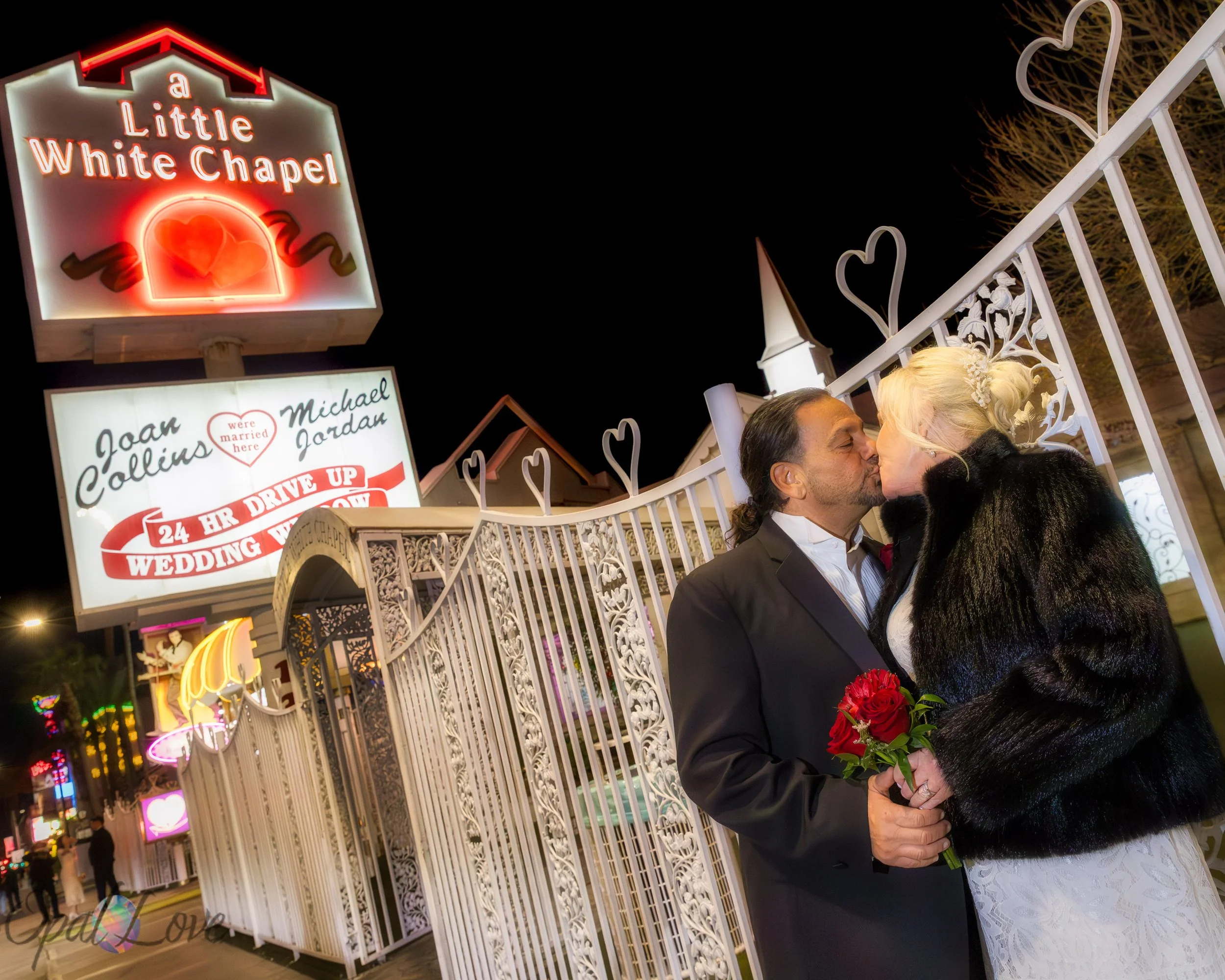 Newlyweds kissing by the white gates and 24 Hour Drive Up Wedding sign outside the Little White Wedding Chapel.