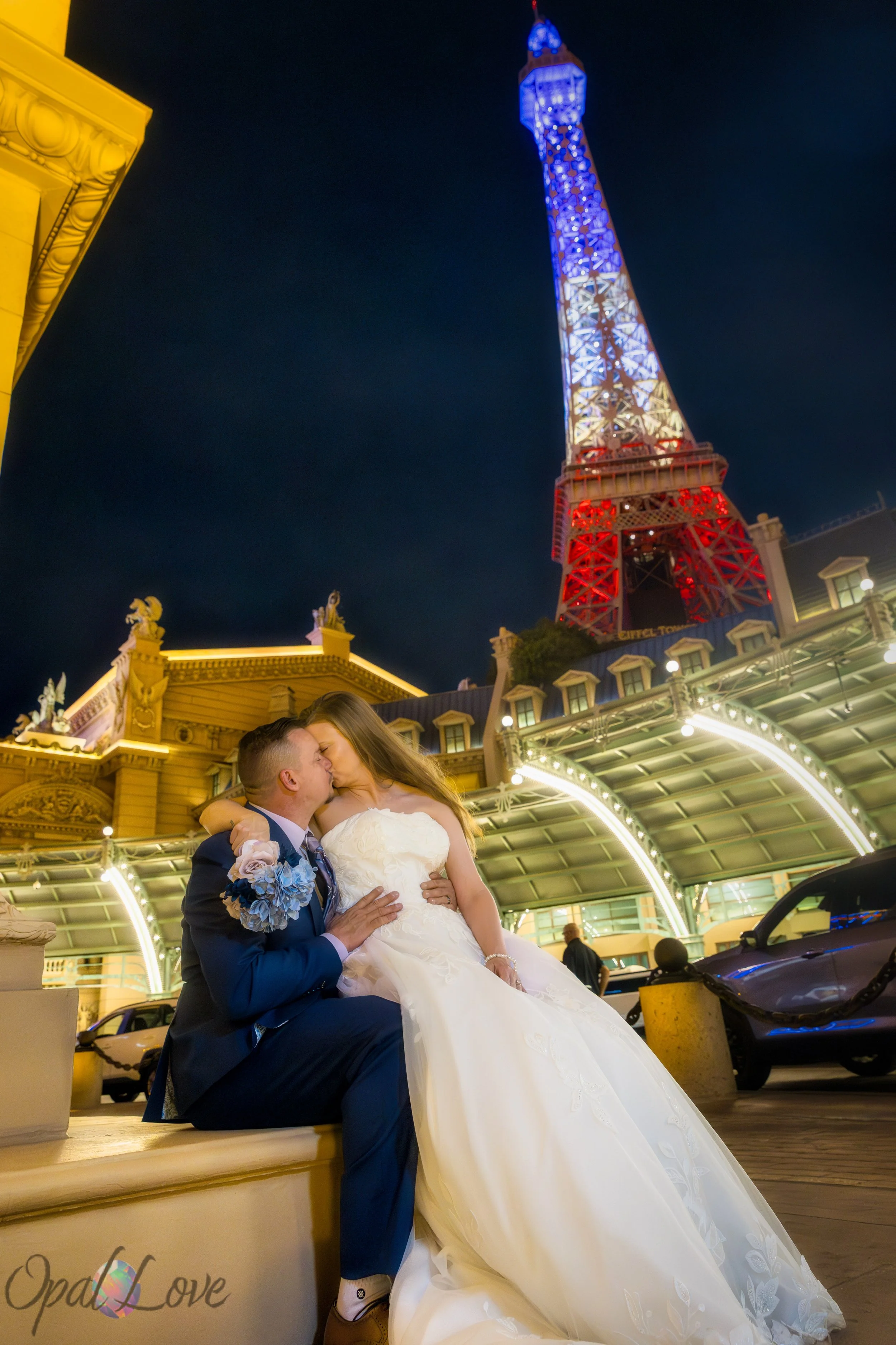 Bride and groom sitting close together sharing a kiss near the Paris Las Vegas entrance at night.