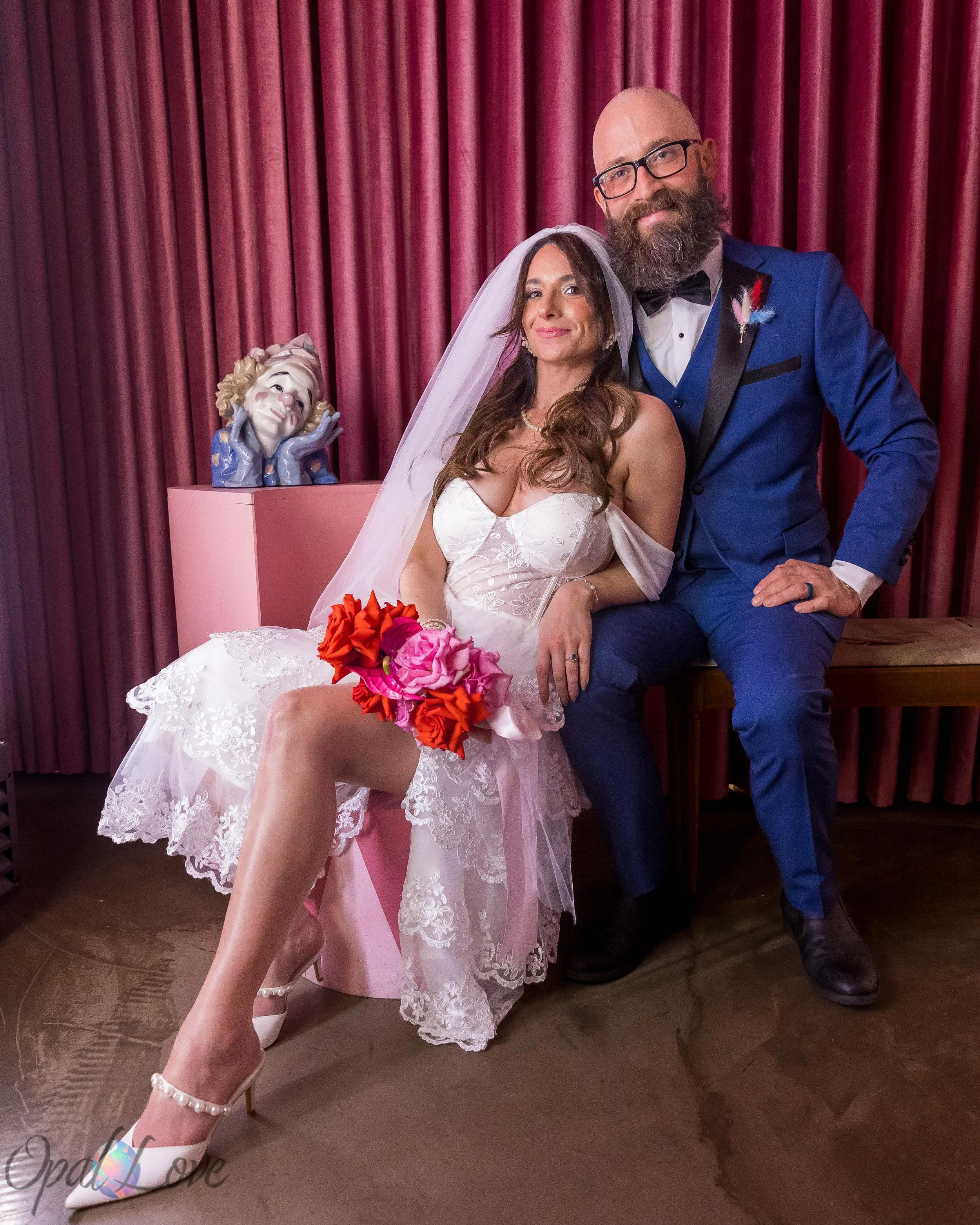 Newlyweds posing together in front of burgundy curtains, with the bride holding a colorful bouquet.