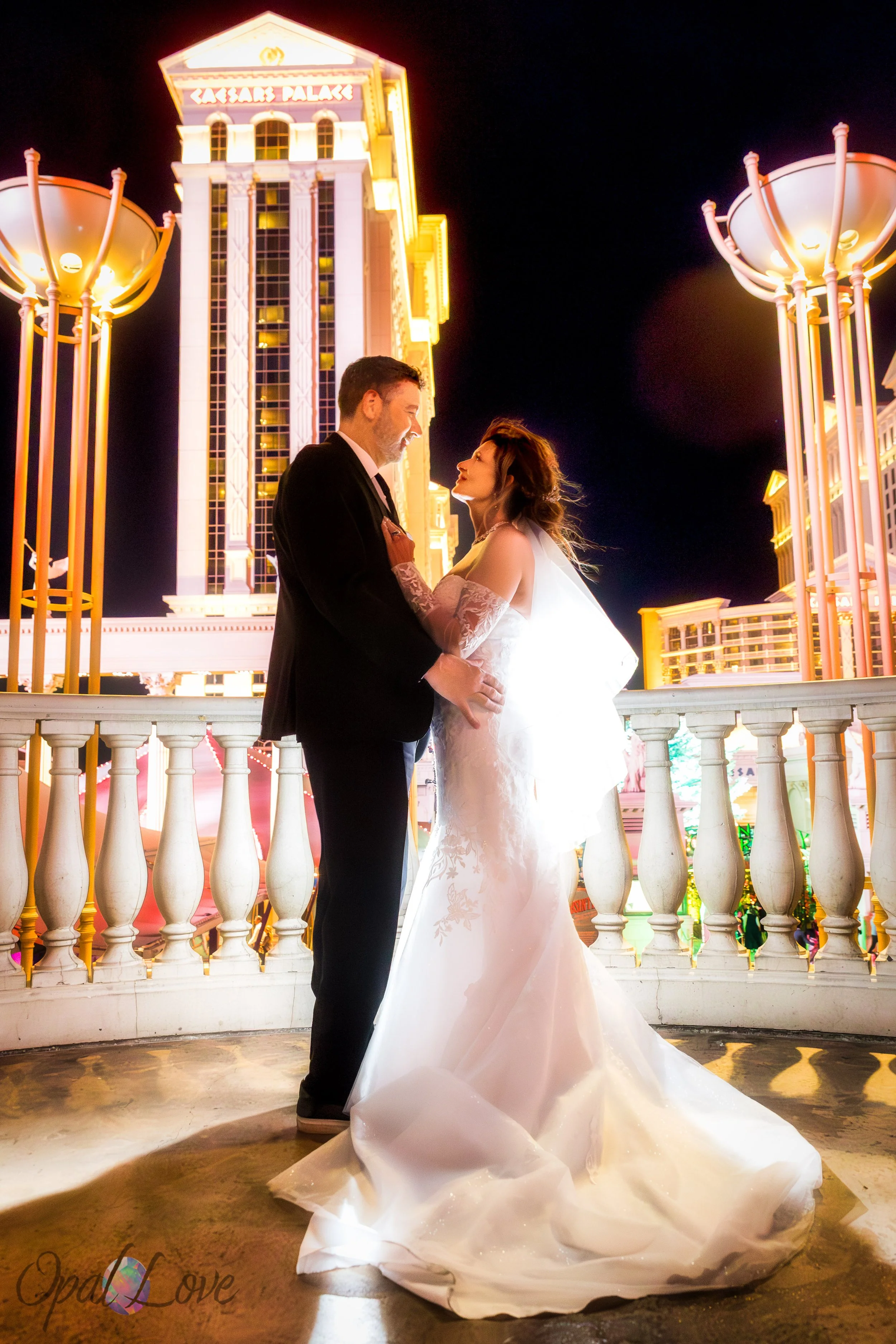 Couple standing on balcony with Caesars Palace tower glowing behind them.