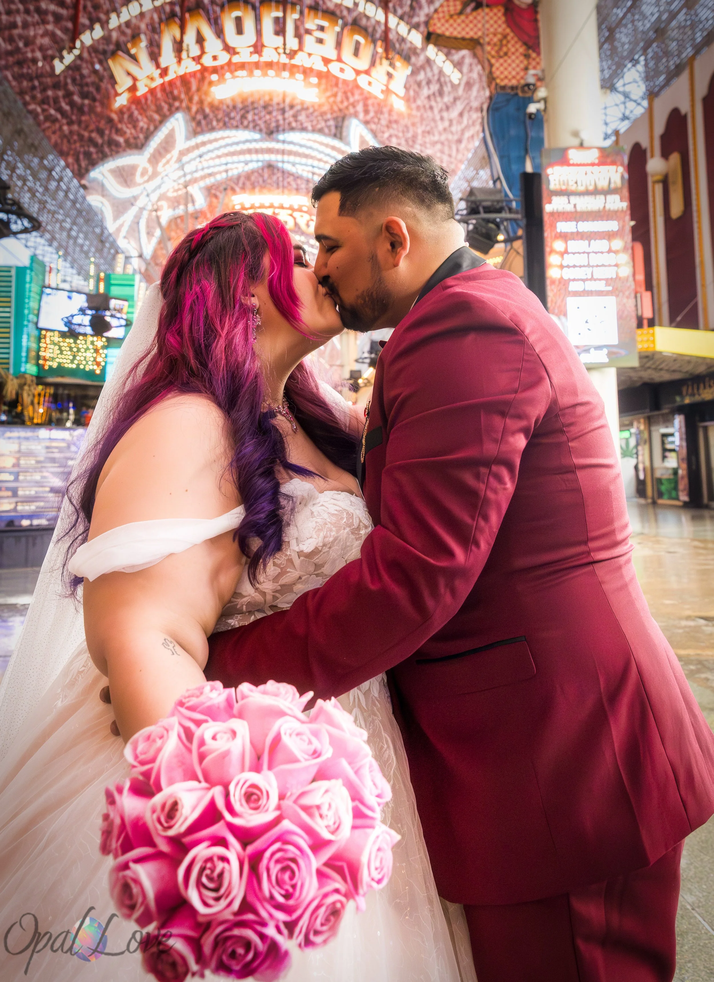 Couple sharing a kiss while the bride holds a bouquet of pink roses toward the camera.