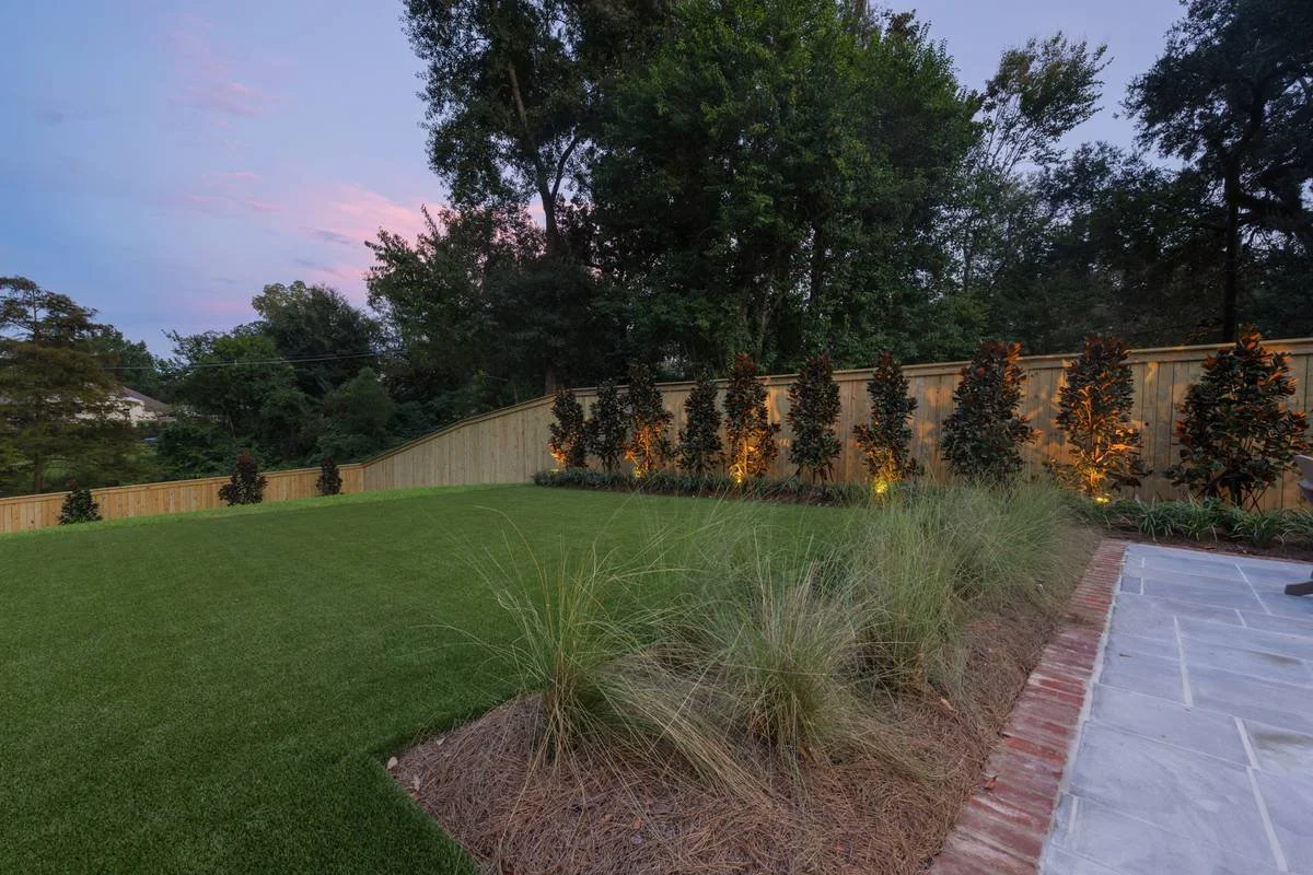 Ornamental grasses and uplit magnolias along the fence at dusk — Flora Landscape Contractors