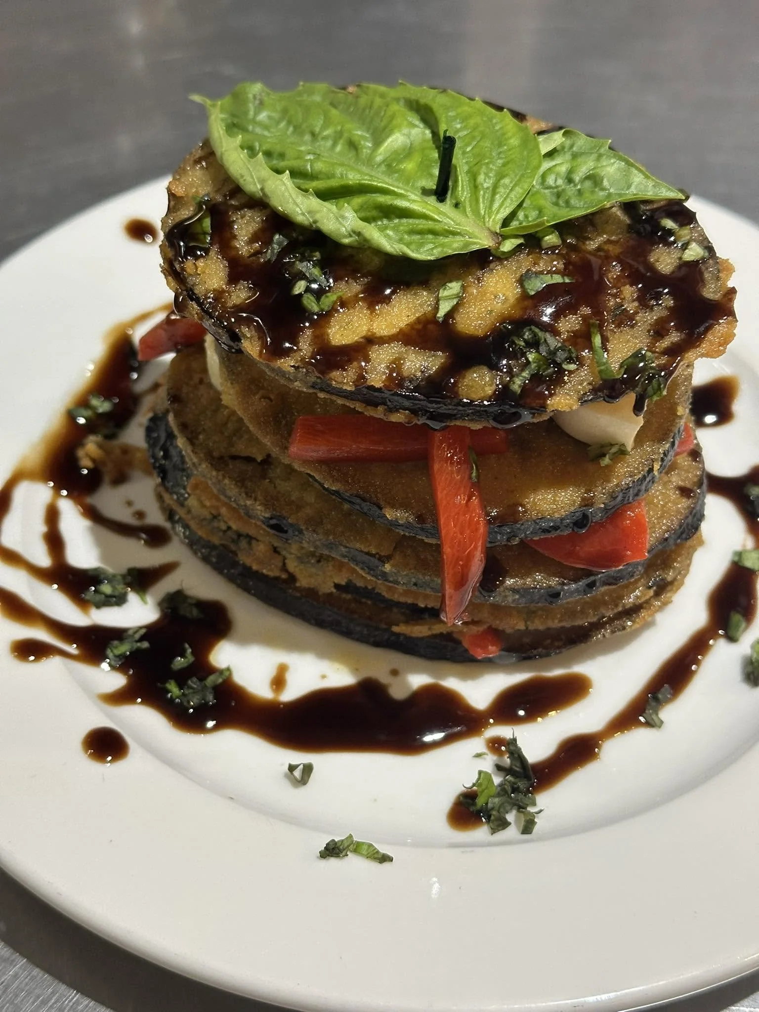 A layered appetizer with slices of fried eggplant, red pepper strips, chopped green herbs, and a green leaf on top, drizzled with dark balsamic glaze and served on a white plate.