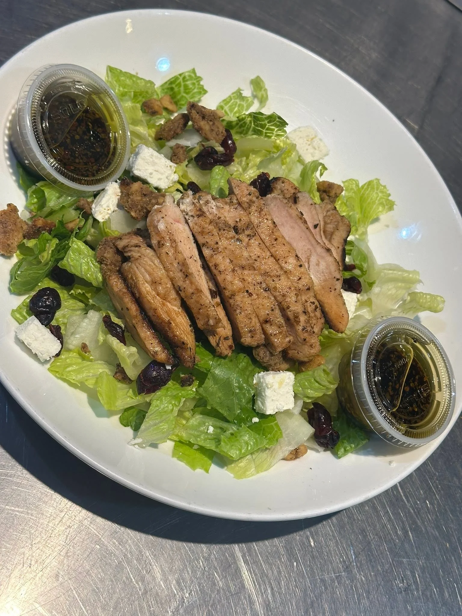 A plate of grilled chicken slices on a bed of green lettuce, topped with crumbled feta cheese, dried cranberries, and croutons, served with two small containers of dressing.