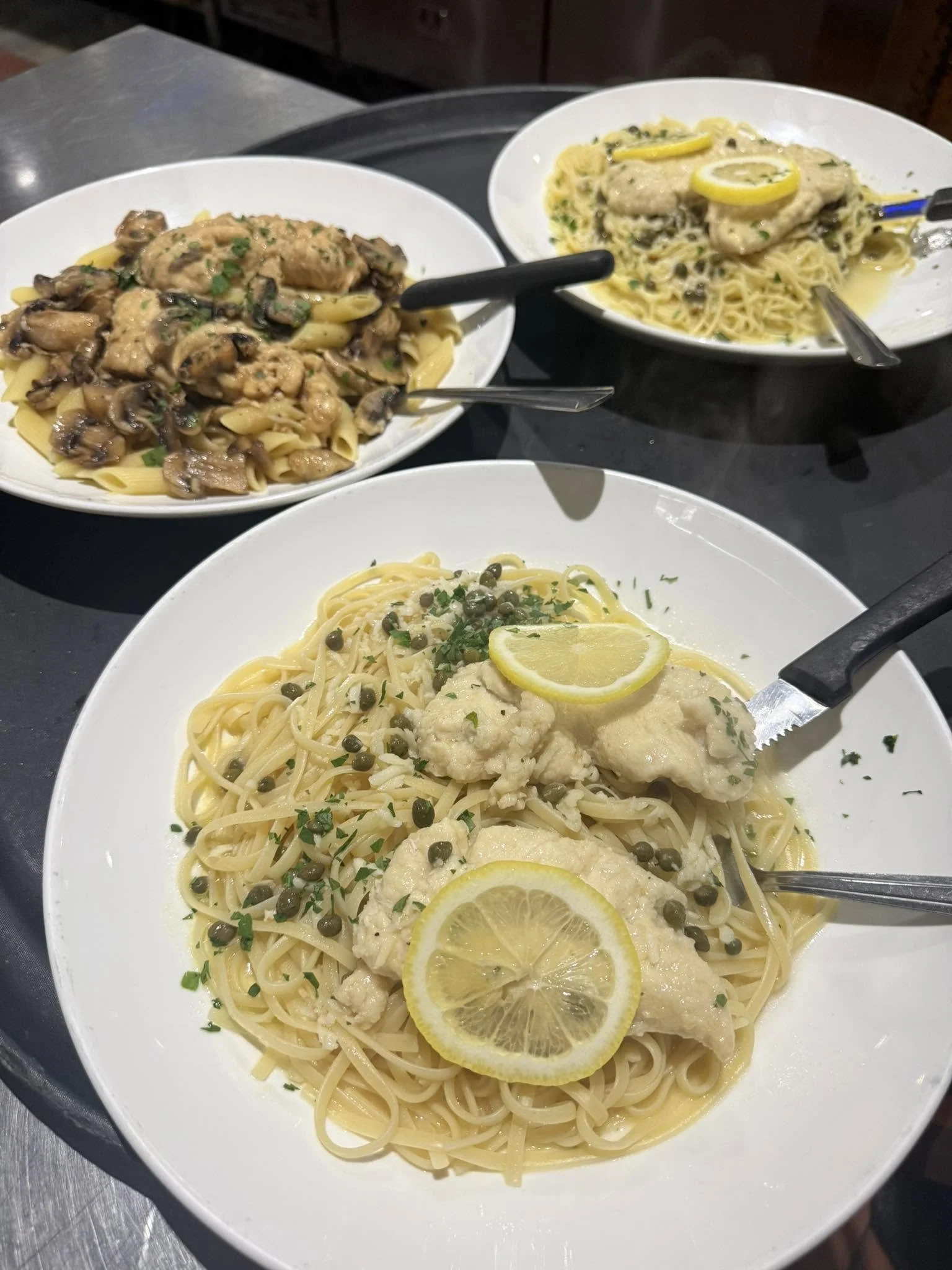 A white plate of spaghetti with lemon slices, topped with green herbs and capers. In the background, two additional bowls contain pasta with chicken and mushrooms, and pasta with chicken in a creamy sauce with lemon slices.