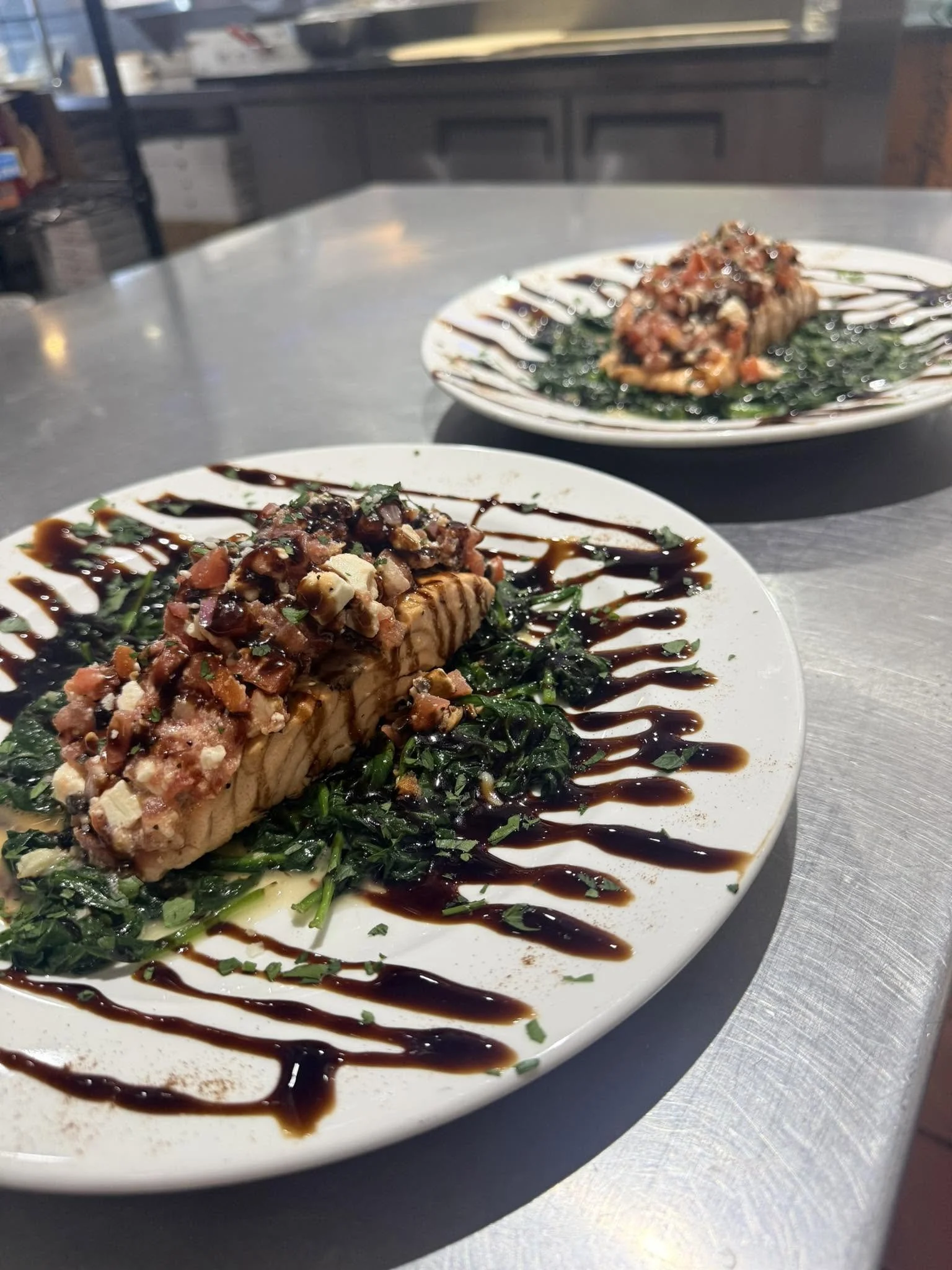 Two plated dishes with seared fish topped with chopped nuts and a tomato-based sauce, garnished with chopped herbs, drizzled with balsamic glaze, and served over cooked greens.