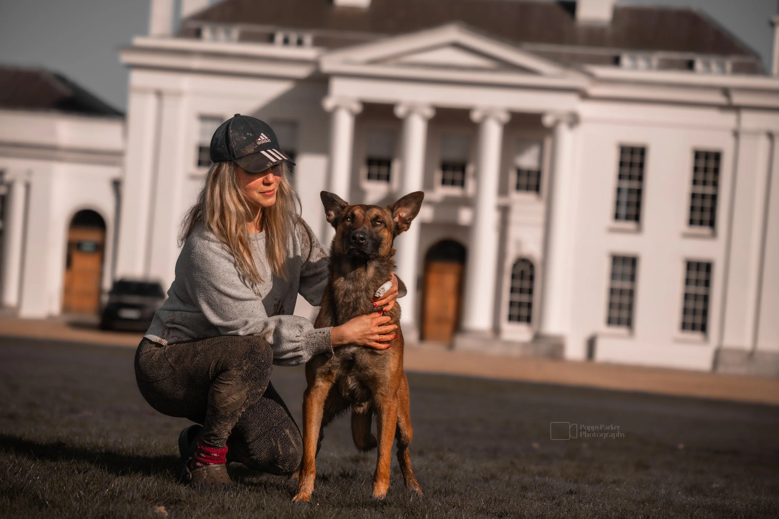female blonde dog trainer with malinois infront of large white home