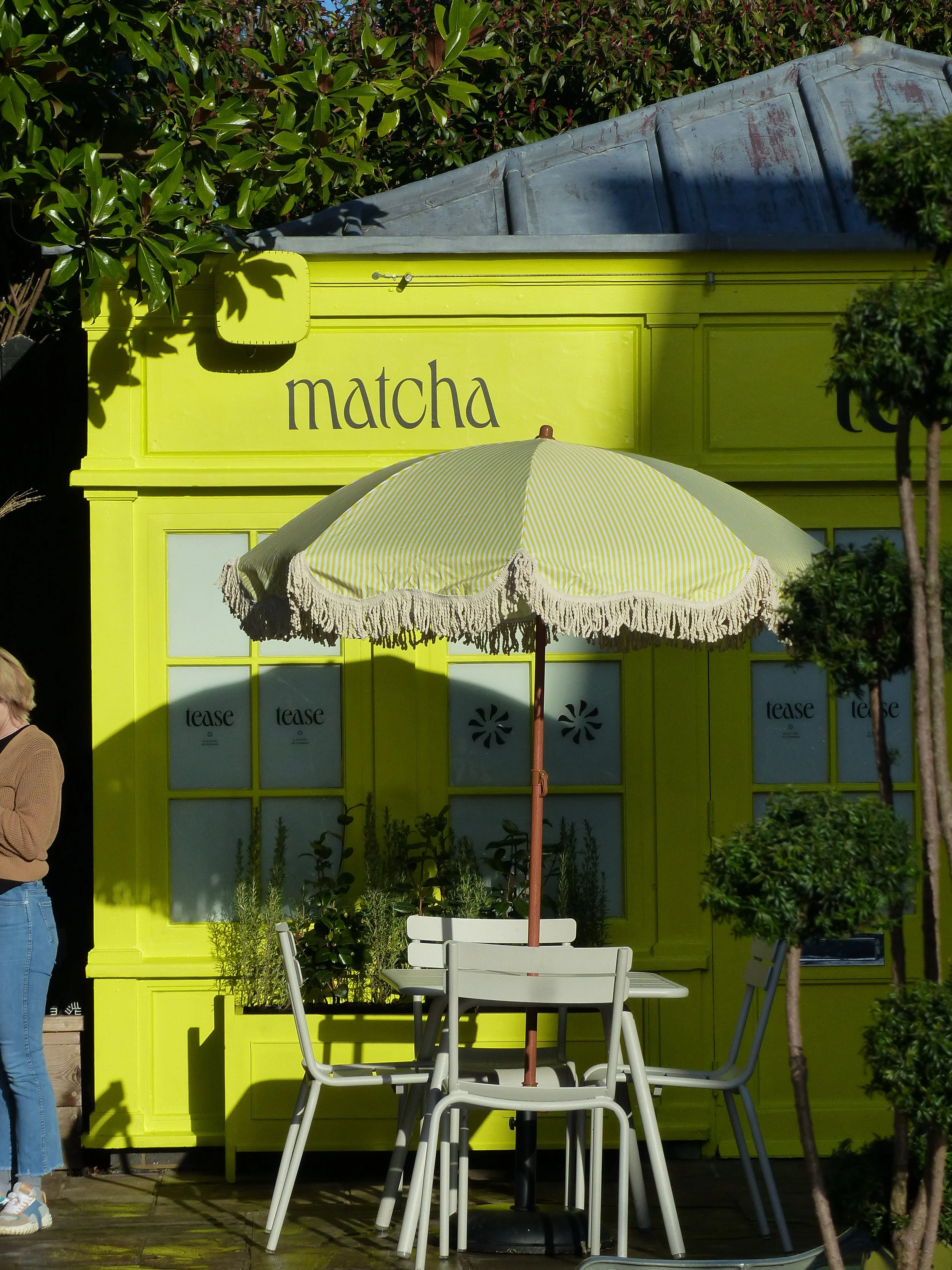 Bright yellow storefront with the word 'matcha' written on it, outdoor seating area with a white table, chairs, and a yellow striped umbrella, surrounded by greenery.