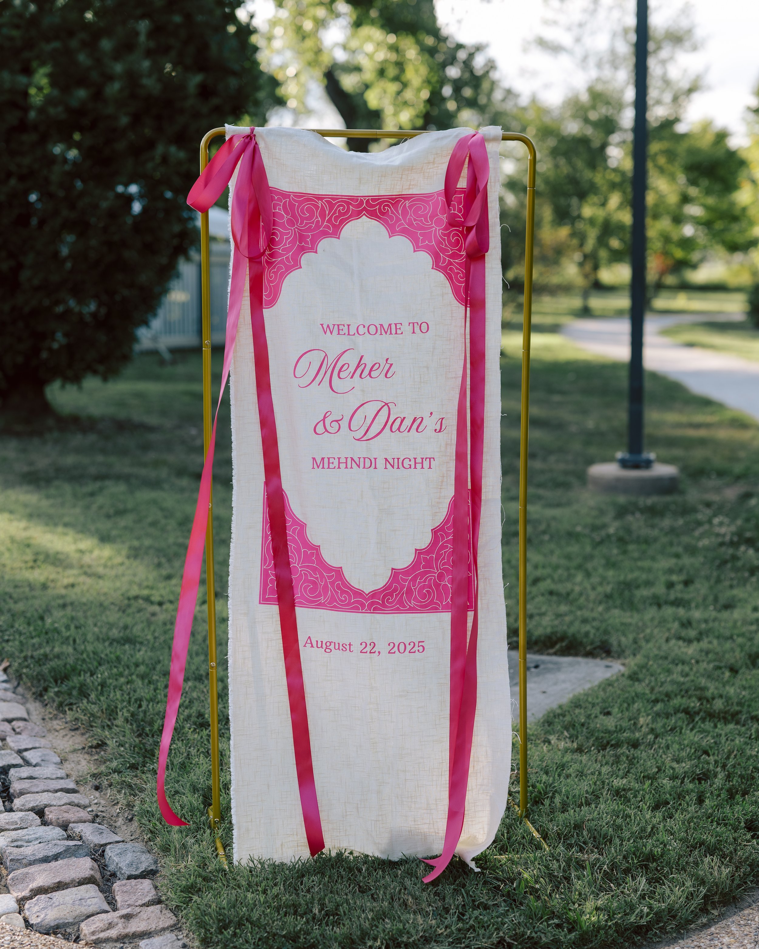 Outdoor welcoming sign on a stand, decorated with pink ribbons, for Meher and Dan's Mehndi Night on August 22, 2025.