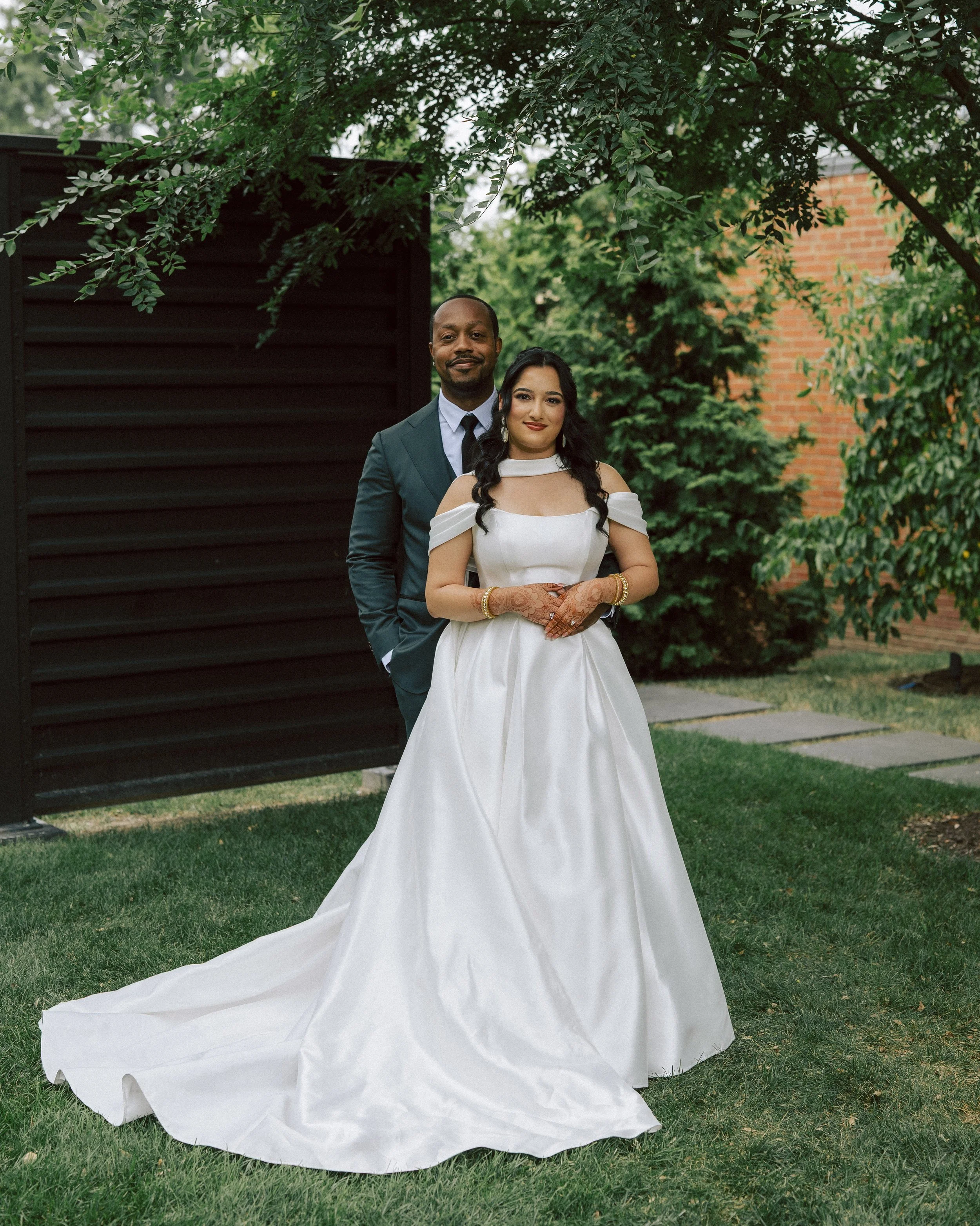 A bride and groom stand together outdoors in a garden setting, with the bride in a white wedding gown and the groom in a dark suit. They are smiling and posing for a photo under a tree.