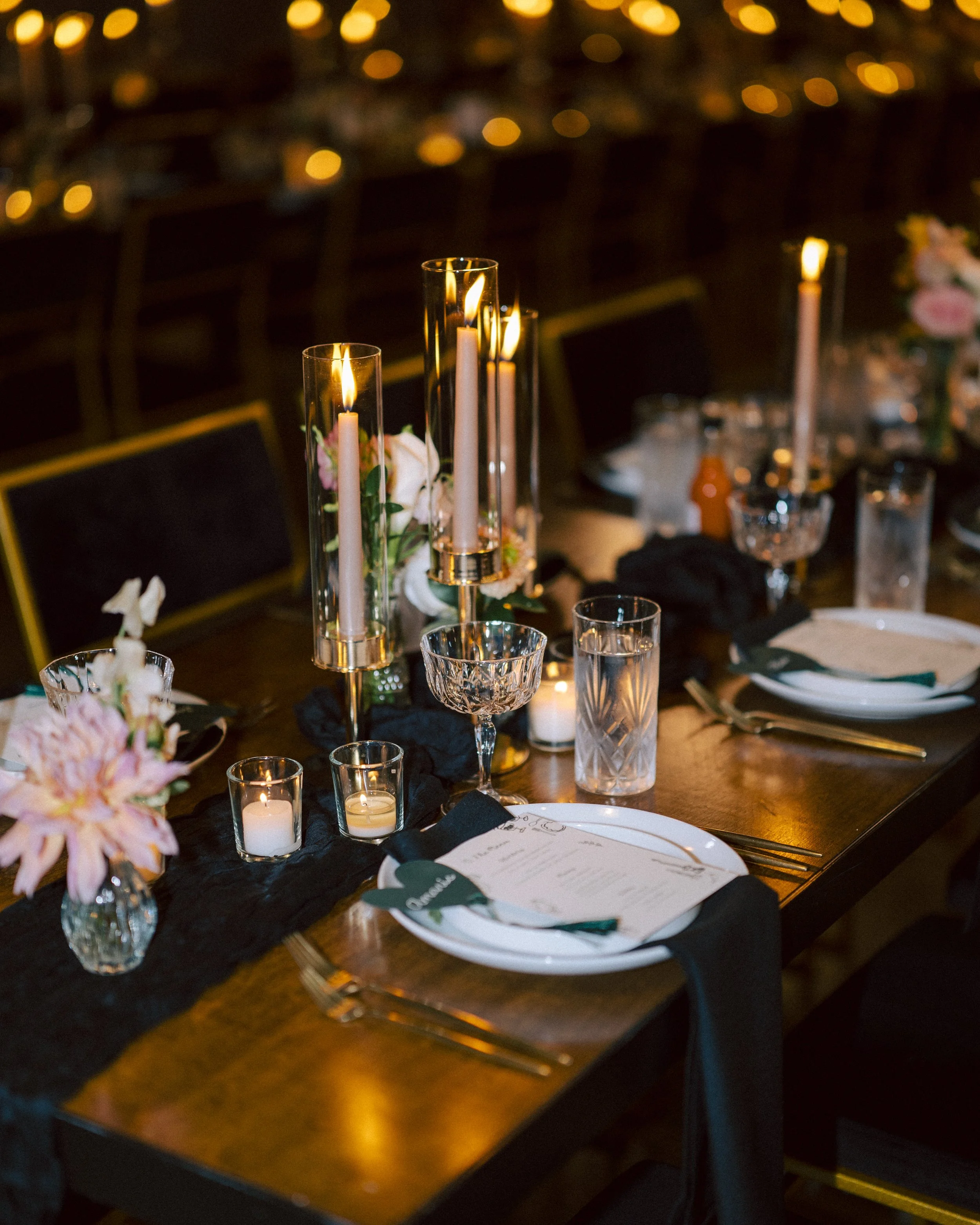 Elegant dining table set for a formal event with tall candles, small votive candles, floral arrangements, glassware, and place settings with menus and napkins.
