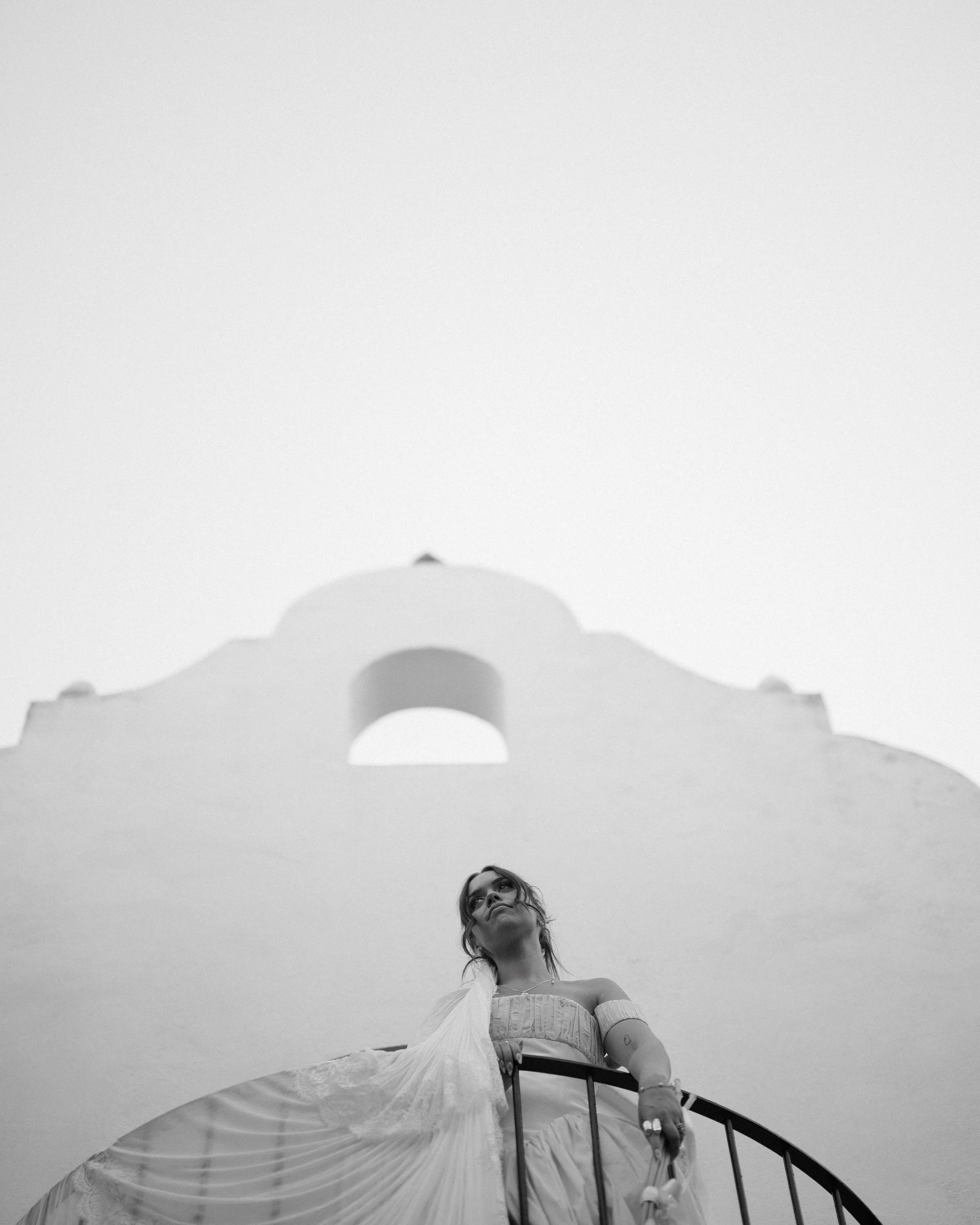 Black and white photo of a woman standing on a curved staircase in front of a white building with a large arch and dome, looking upward.