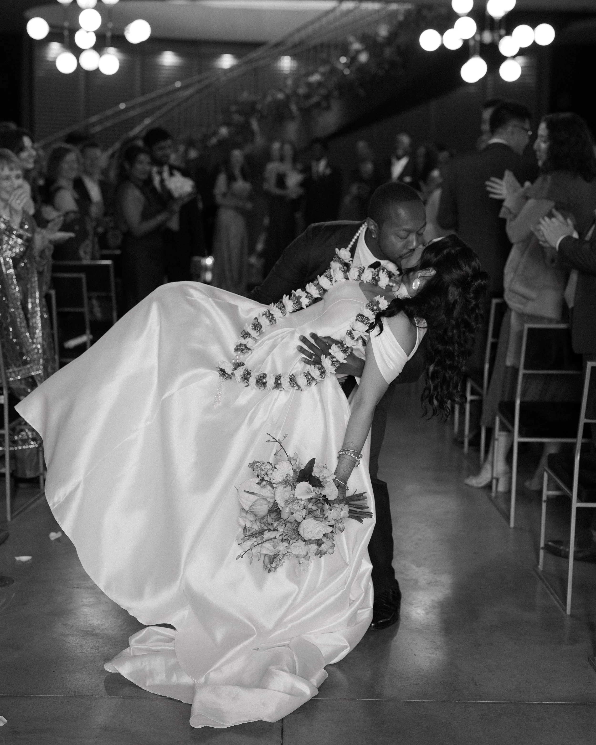 A black and white photo of a newlywed couple at their wedding reception, with the groom dipping the bride for a kiss while she holds a bouquet of flowers, surrounded by wedding guests.