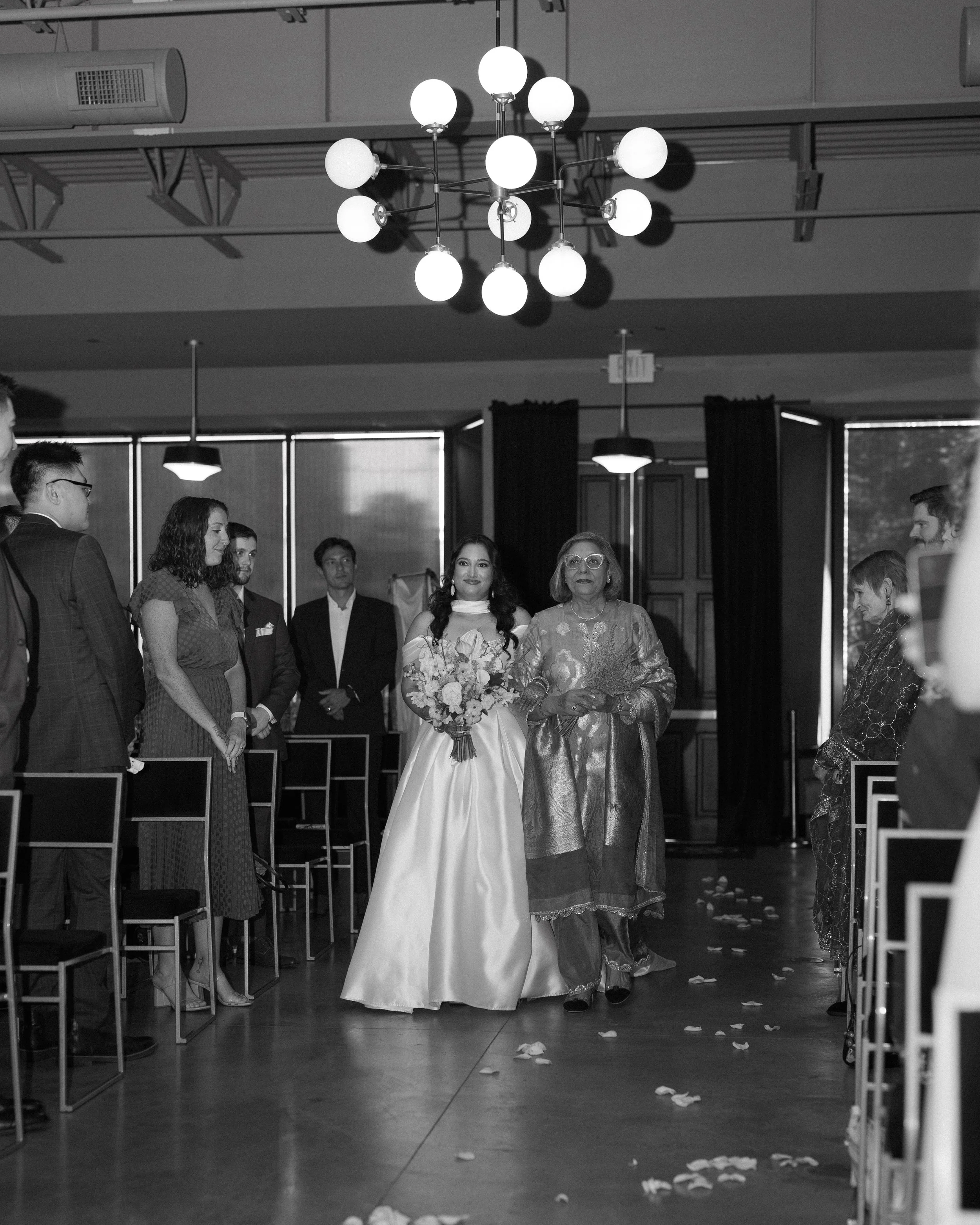 A bride walking down the aisle with an older woman at a wedding ceremony in a decorated indoor venue.