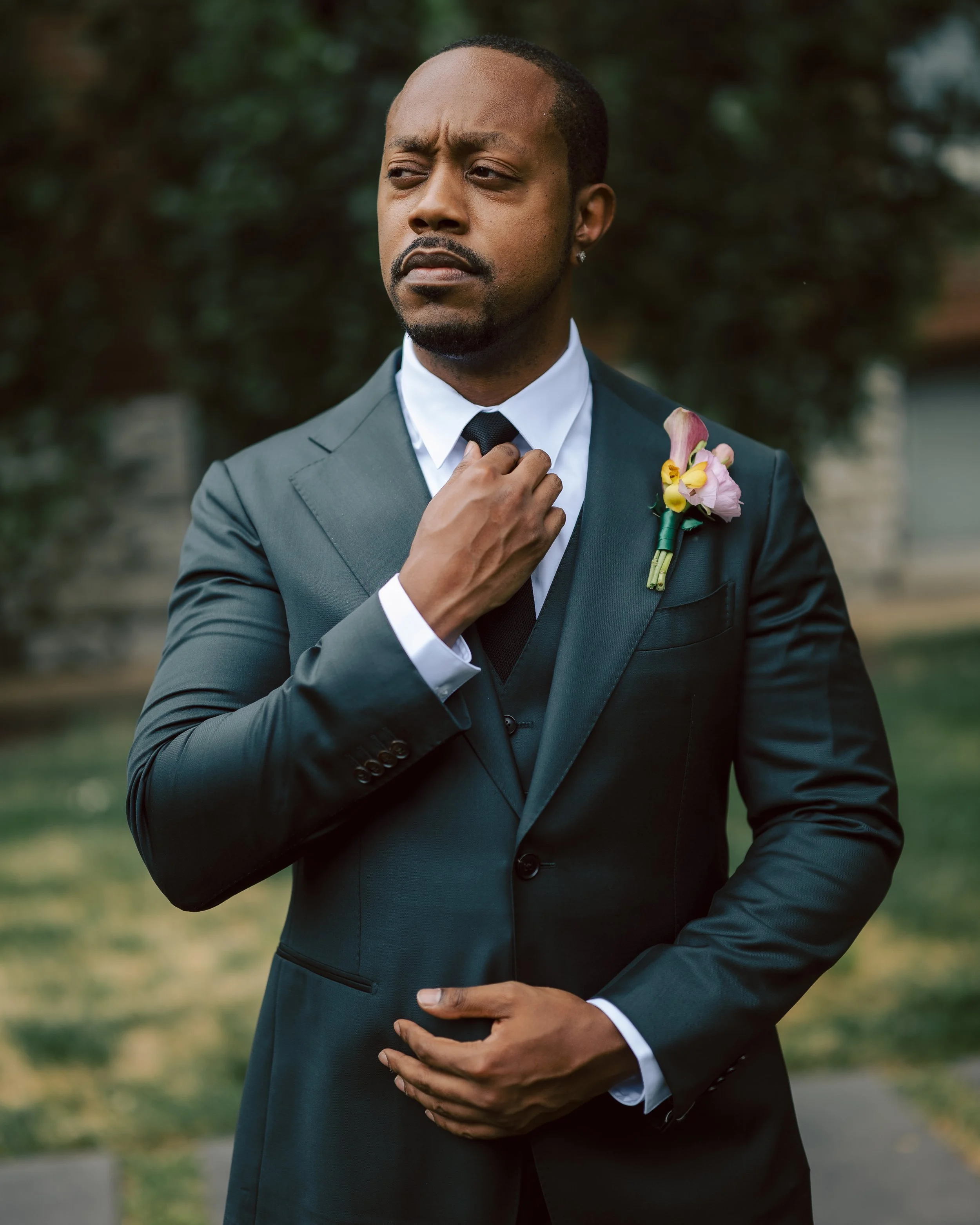 A man in a black suit and tie, adjusting his tie, with a boutonnière on his lapel, standing outdoors in a garden or park.