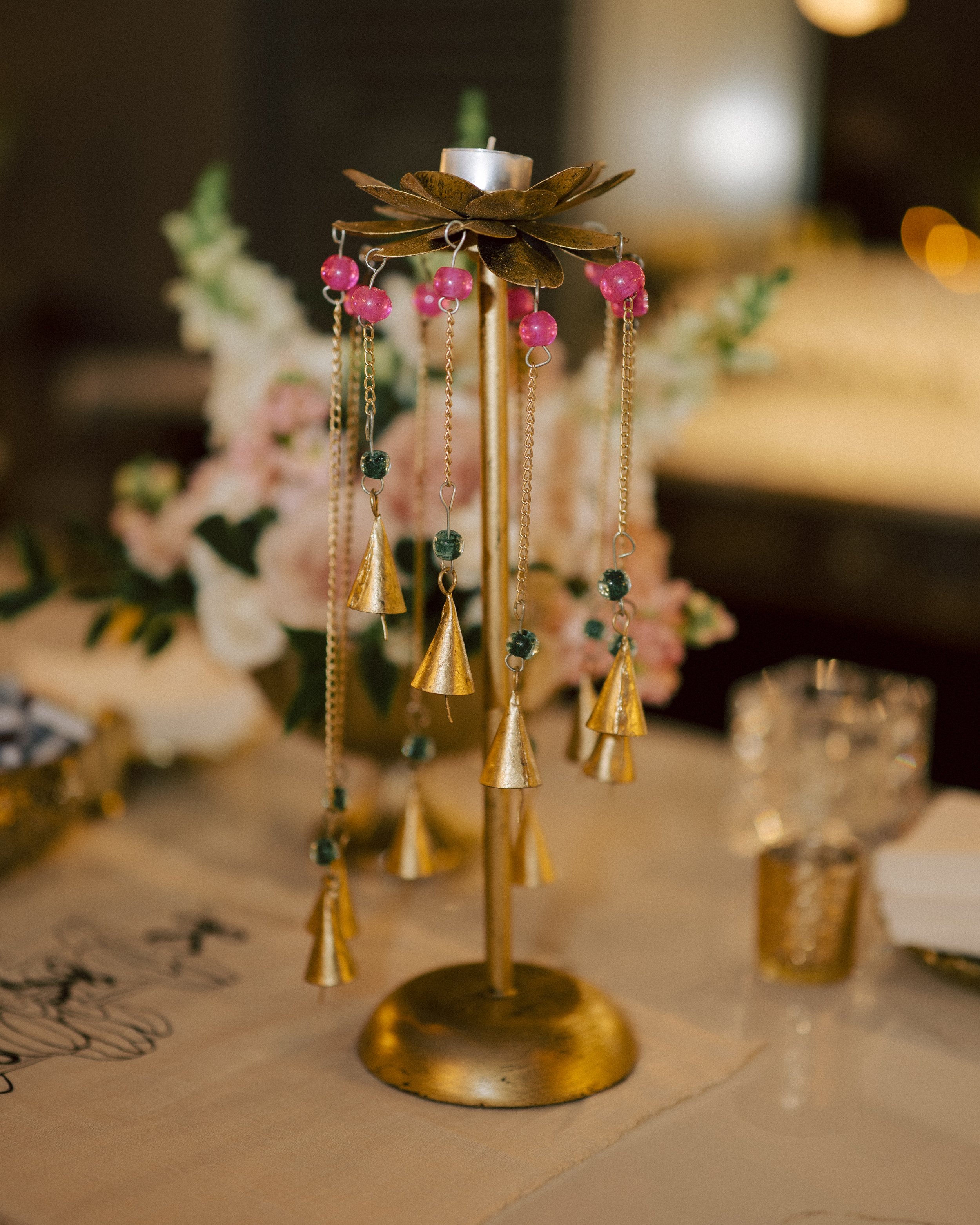Gold jewelry stand holding pink and green beaded earrings, with flowers and table setting in the background.