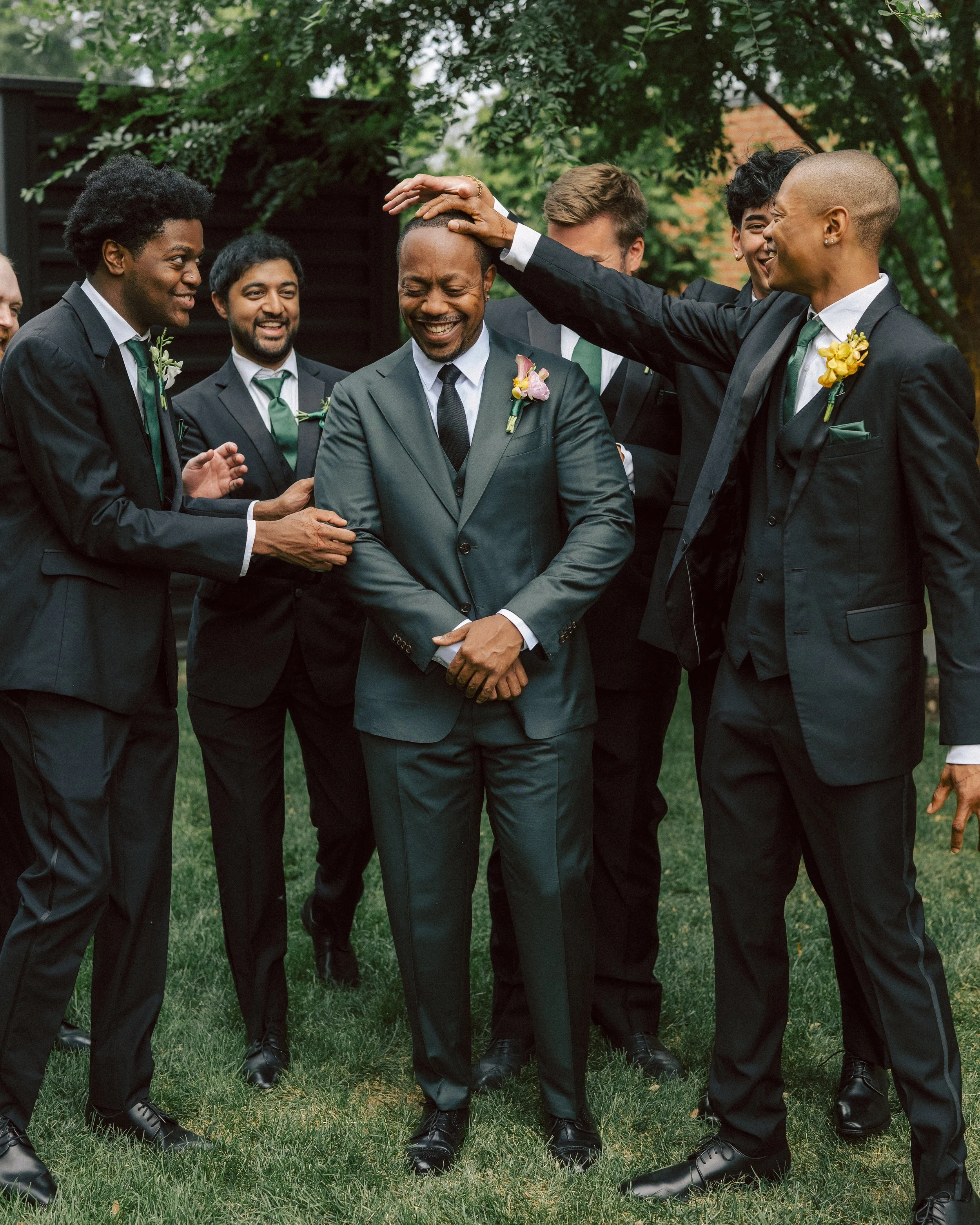 Group of men in suits celebrating outdoors, with one man in the center laughing and others touching his head