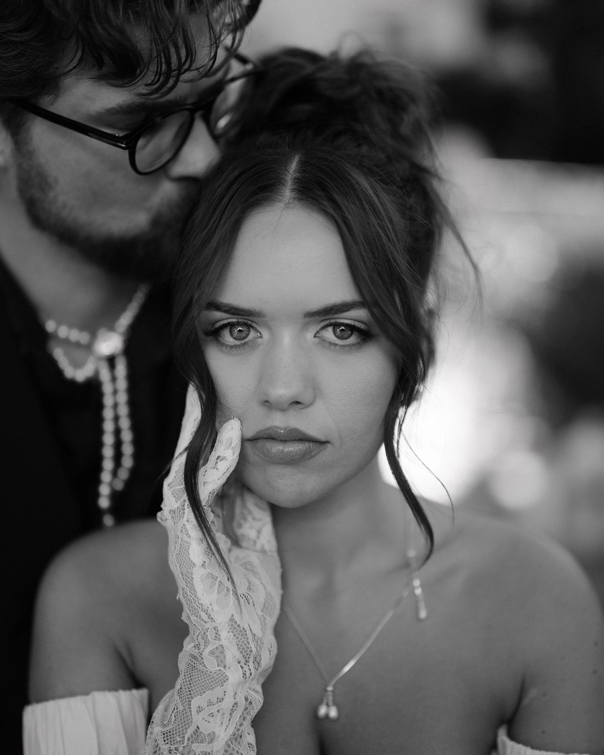 A black-and-white photo of a man with glasses and a woman with wavy hair. The man is kissing the woman's head while she looks directly at the camera.