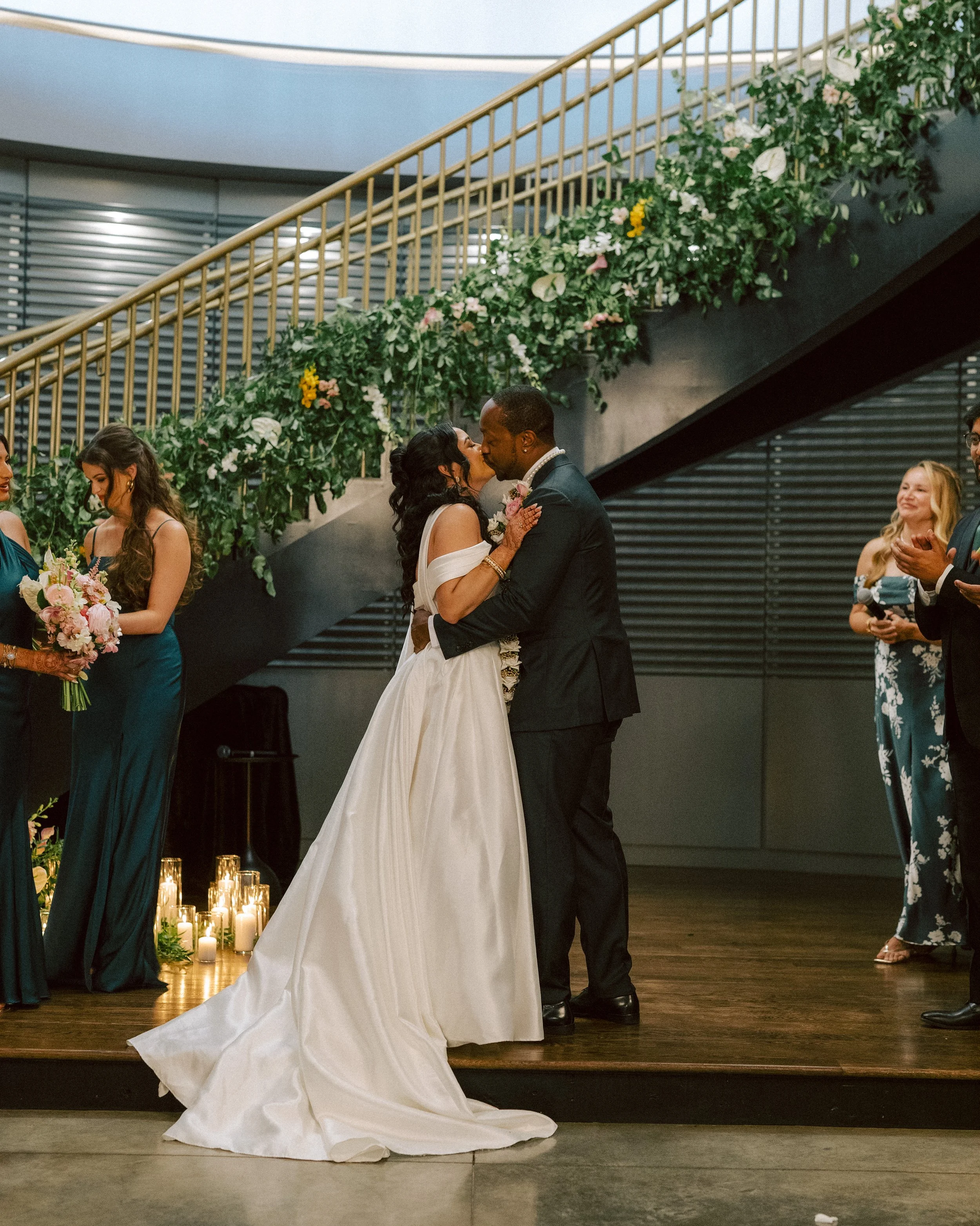 A bride and groom kiss during their wedding ceremony, standing on a wooden platform with candles at their feet, surrounded by bridesmaids and guests, with a staircase decorated with flowers in the background.