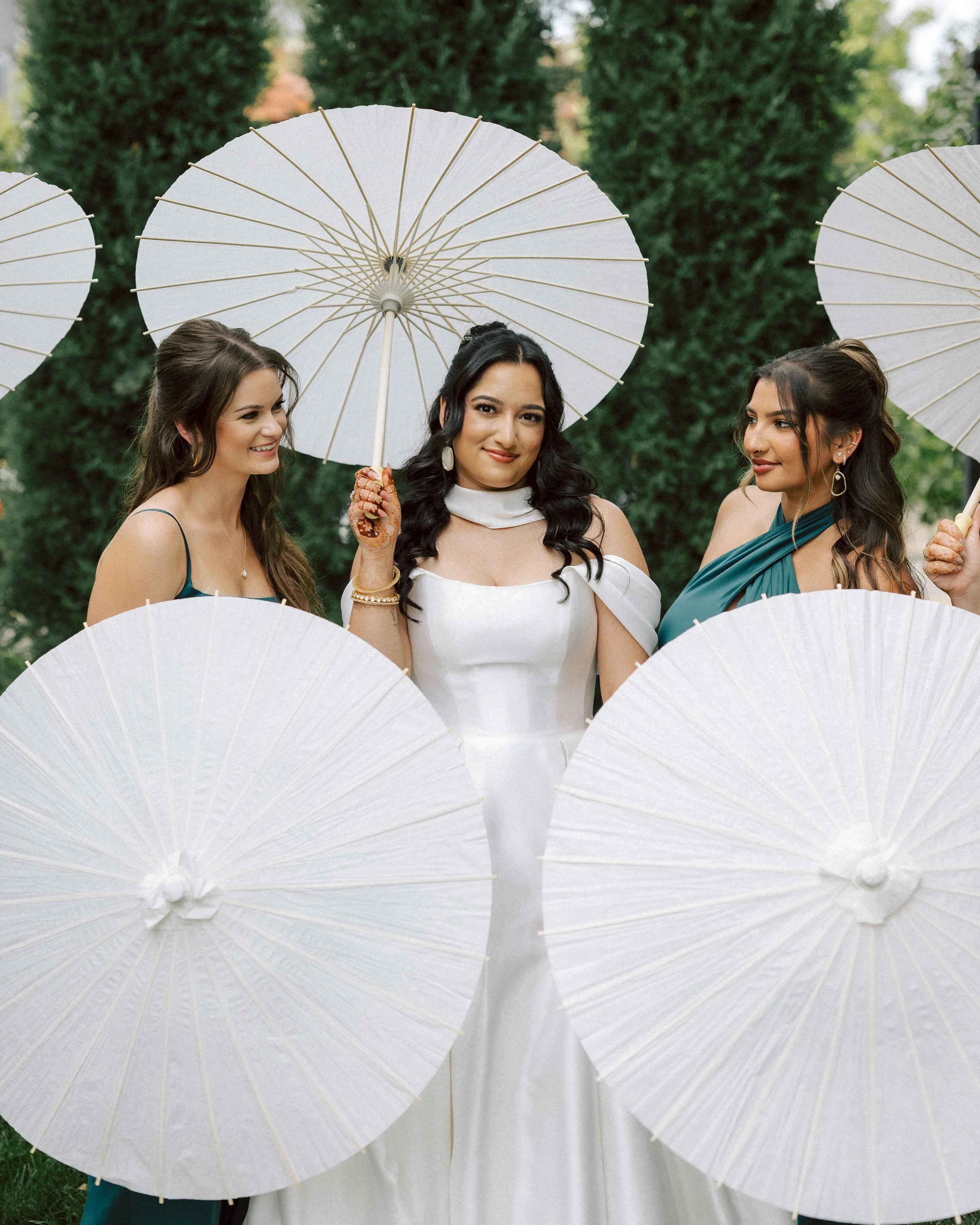 Three women in dresses standing outdoors, holding white parasols, with a background of green trees.