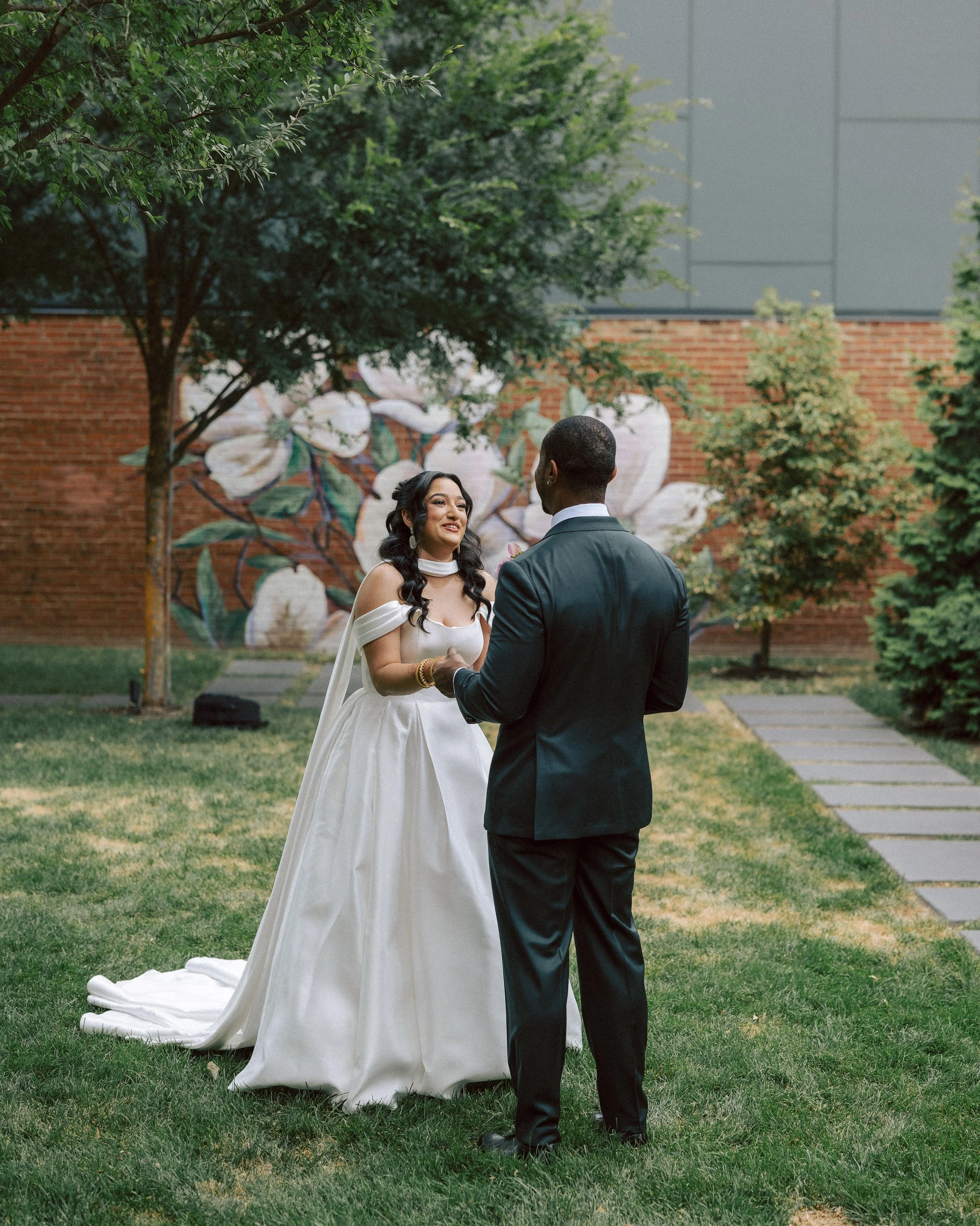 A bride and groom standing in a garden, holding hands and exchanging vows during a wedding ceremony, with trees and a mural on a brick wall in the background.