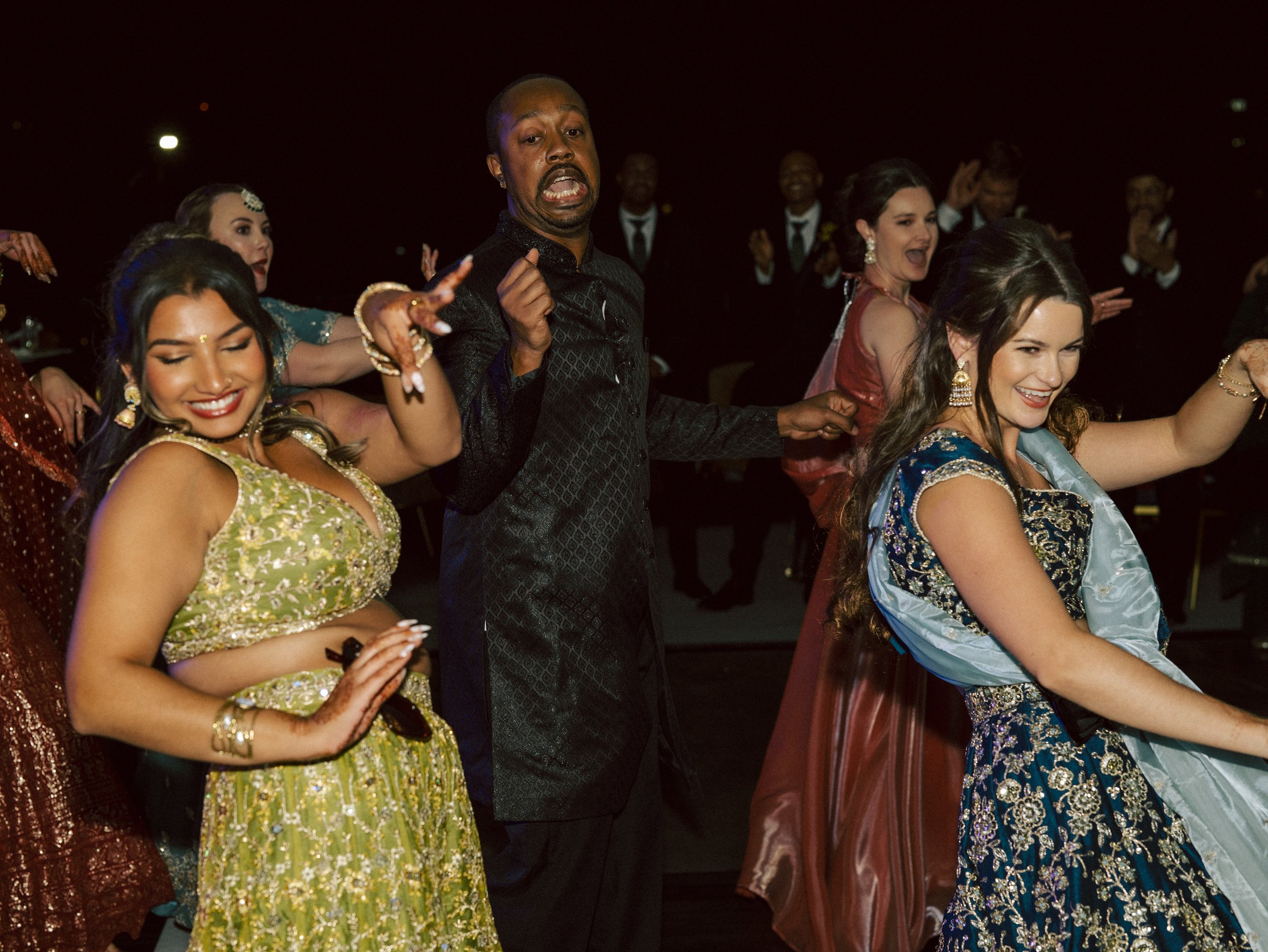 People dressed in traditional Indian attire dancing at a celebration, with a woman in yellow and another in blue, all smiling and enjoying themselves.