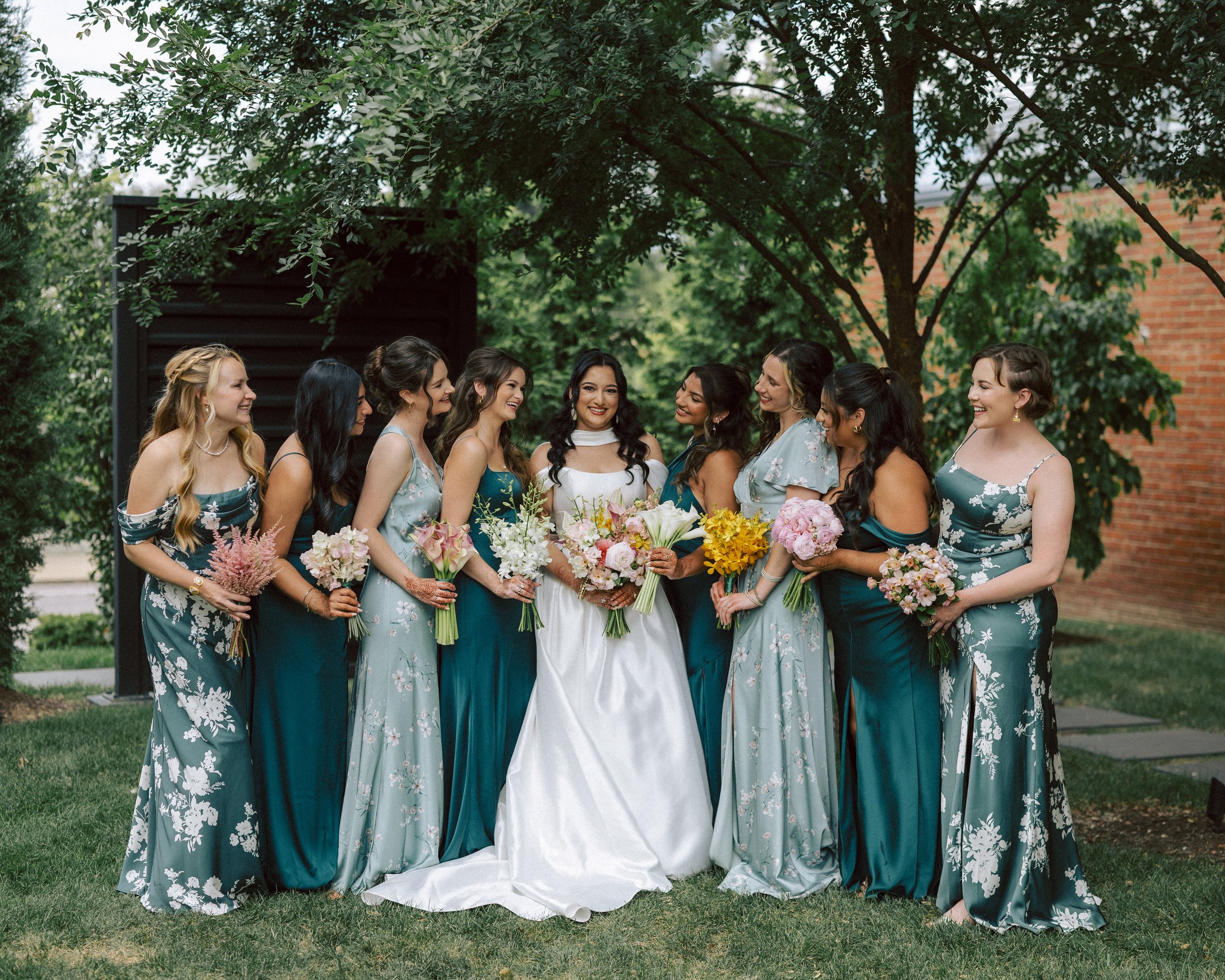 A bride and ten bridesmaids standing outdoors under a tree in garden setting, all holding bouquets of flowers, dressed in formal dresses, smiling and celebrating.
