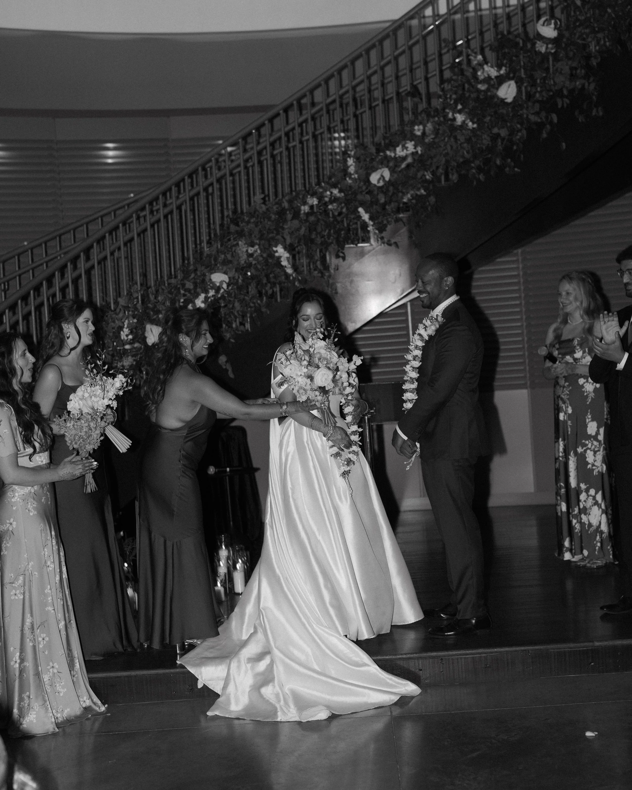 Black and white photo of a wedding ceremony with the bride, groom, and bridesmaids exchanging rings under a floral arch.