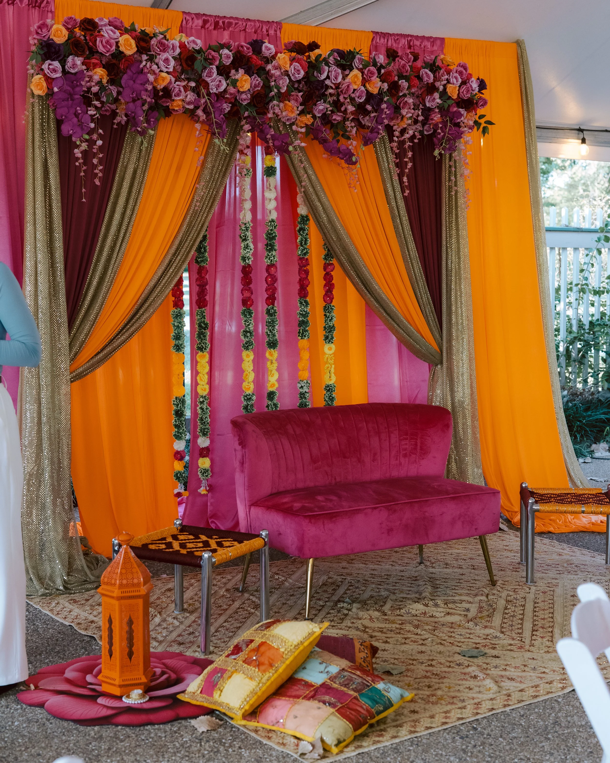 Colorful event setup with pink, orange, gold, and burgundy drapes, floral decorations, a pink velvet loveseat, decorative lantern, cushions, and a patterned rug.