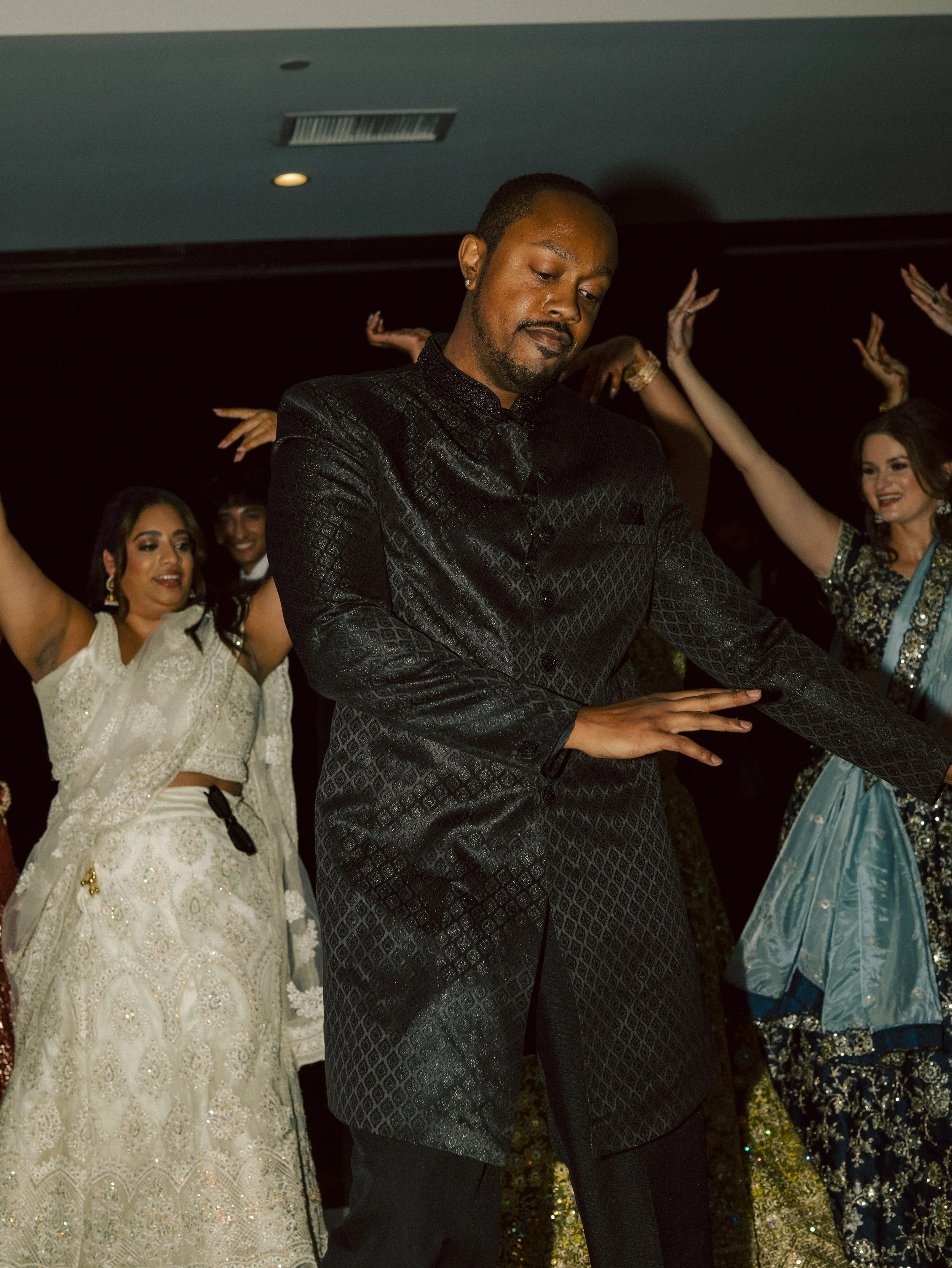 A man in a black traditional Indian outfit dancing with women in colorful Indian attire at a celebration.