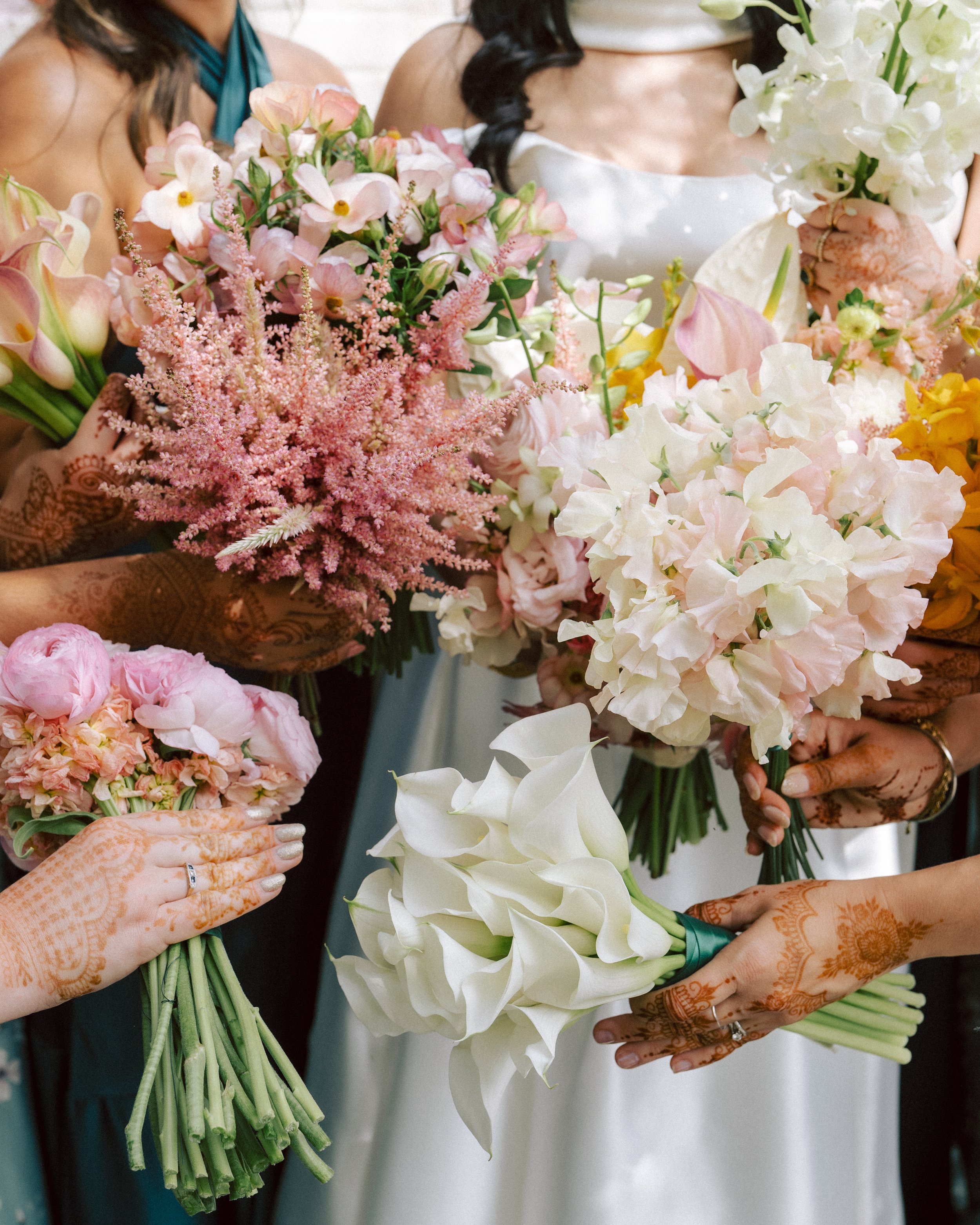Several women holding bouquets of various flowers including white calla lilies, pink and white peonies, and other pink, white, and yellow blooms at a wedding or celebration.