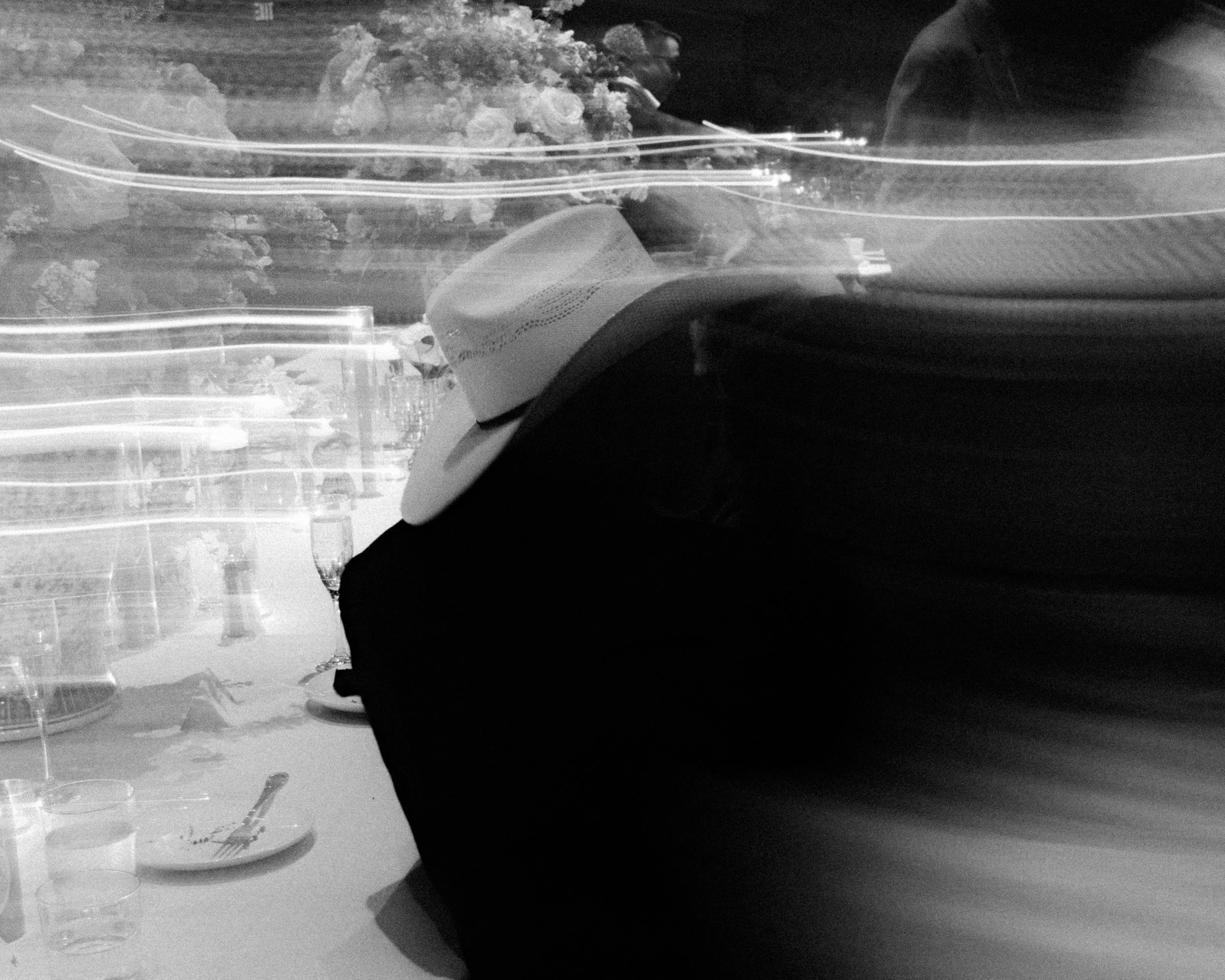 Black and white photo of a man wearing a cowboy hat, seen through a window with light streaks, tables with plates and glasses outside, and some trees in the background.