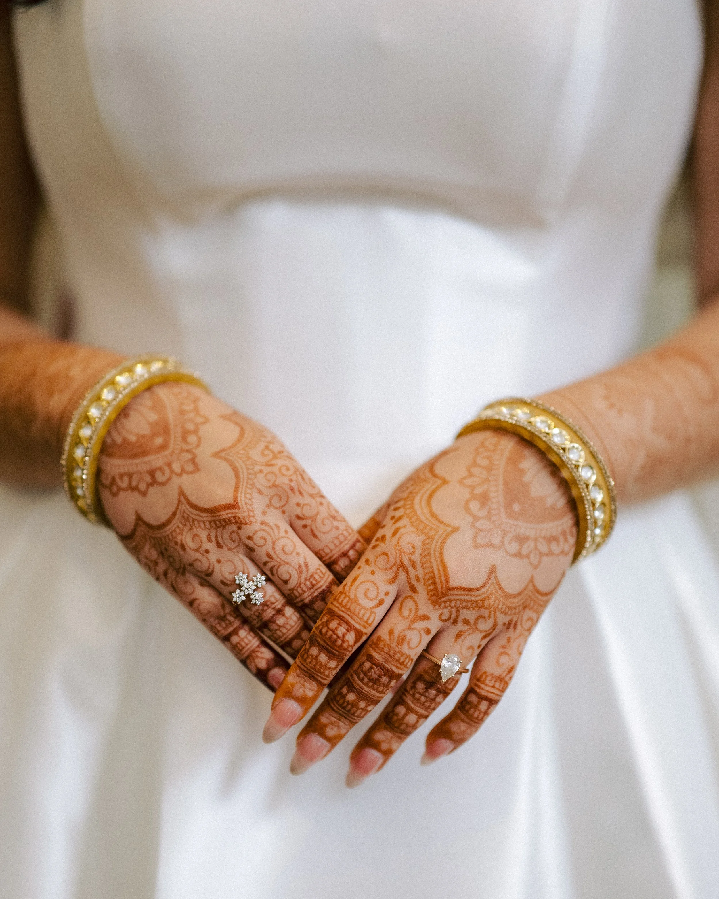 Close-up of a bride's hands with intricate henna designs, gold jewelry, and engagement rings, set against a white dress.