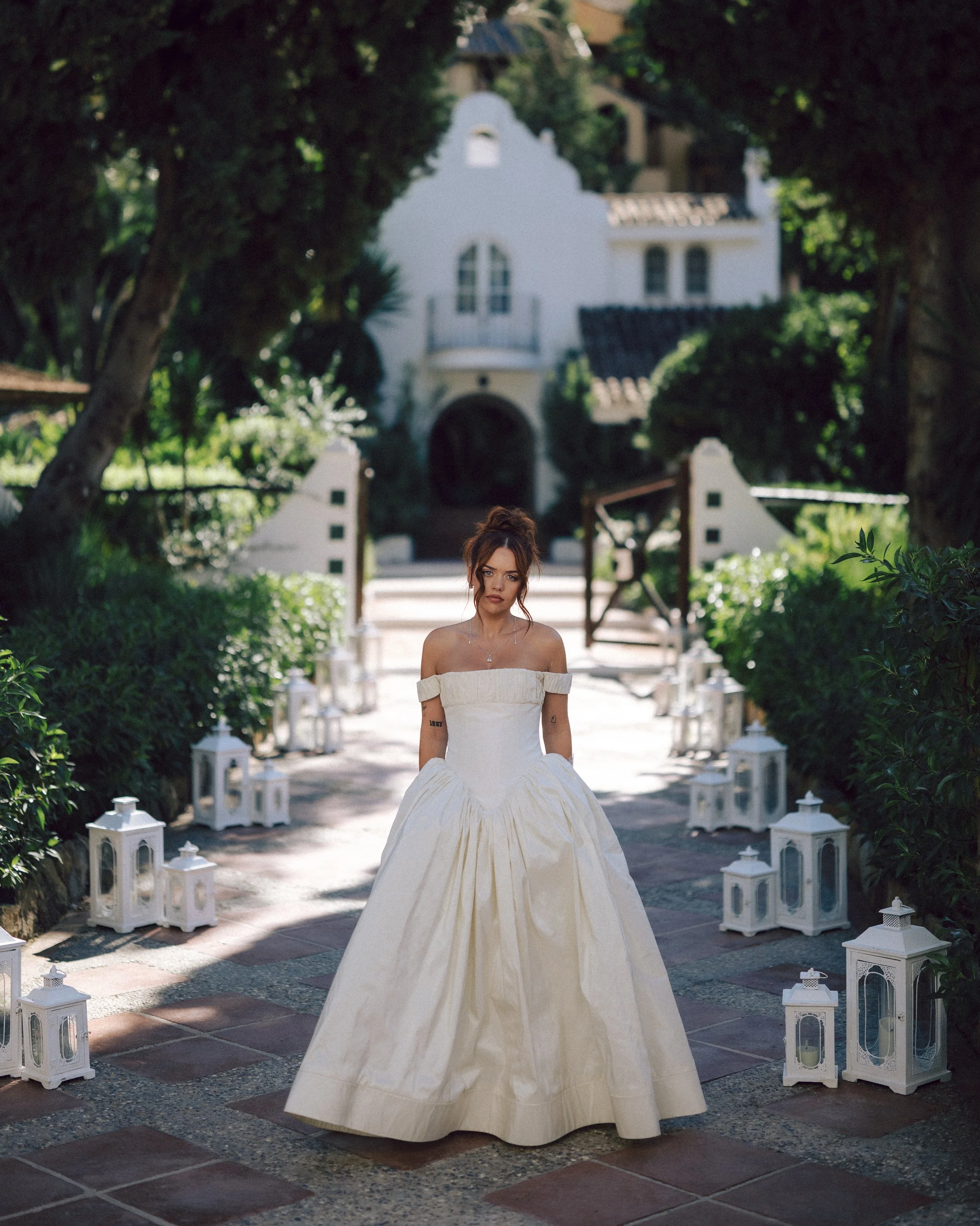 A woman in a white off-shoulder vintage wedding dress standing on a pathway lined with white lanterns, with a large white house and greenery in the background.