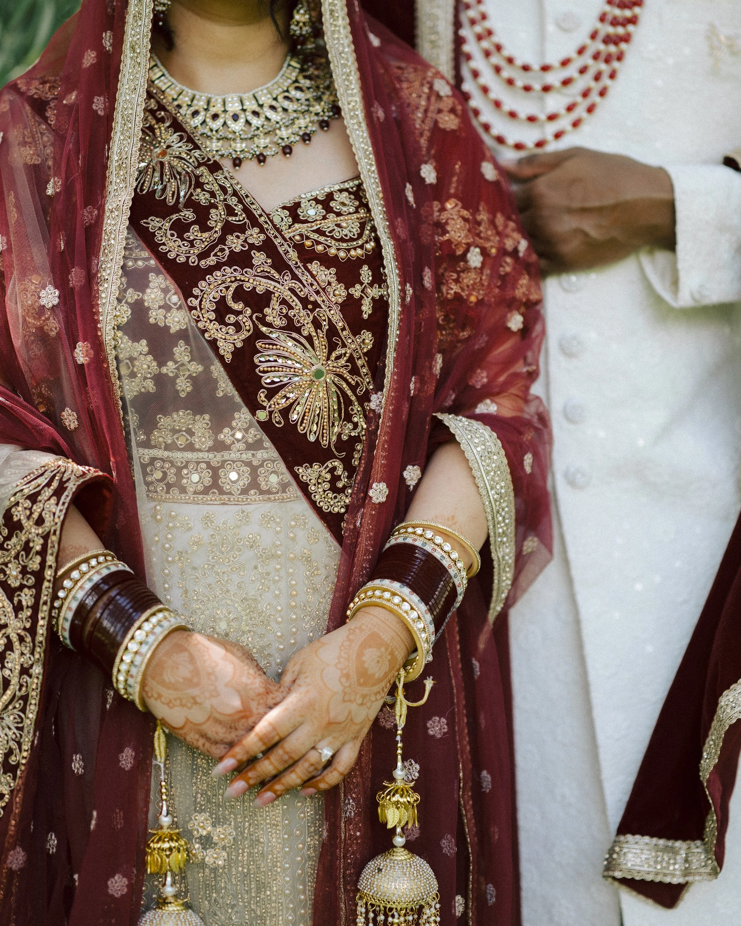 Close-up of a bride wearing a red and gold traditional Indian outfit with intricate embroidery, jewelry, and mehndi on her hands during a wedding ceremony.