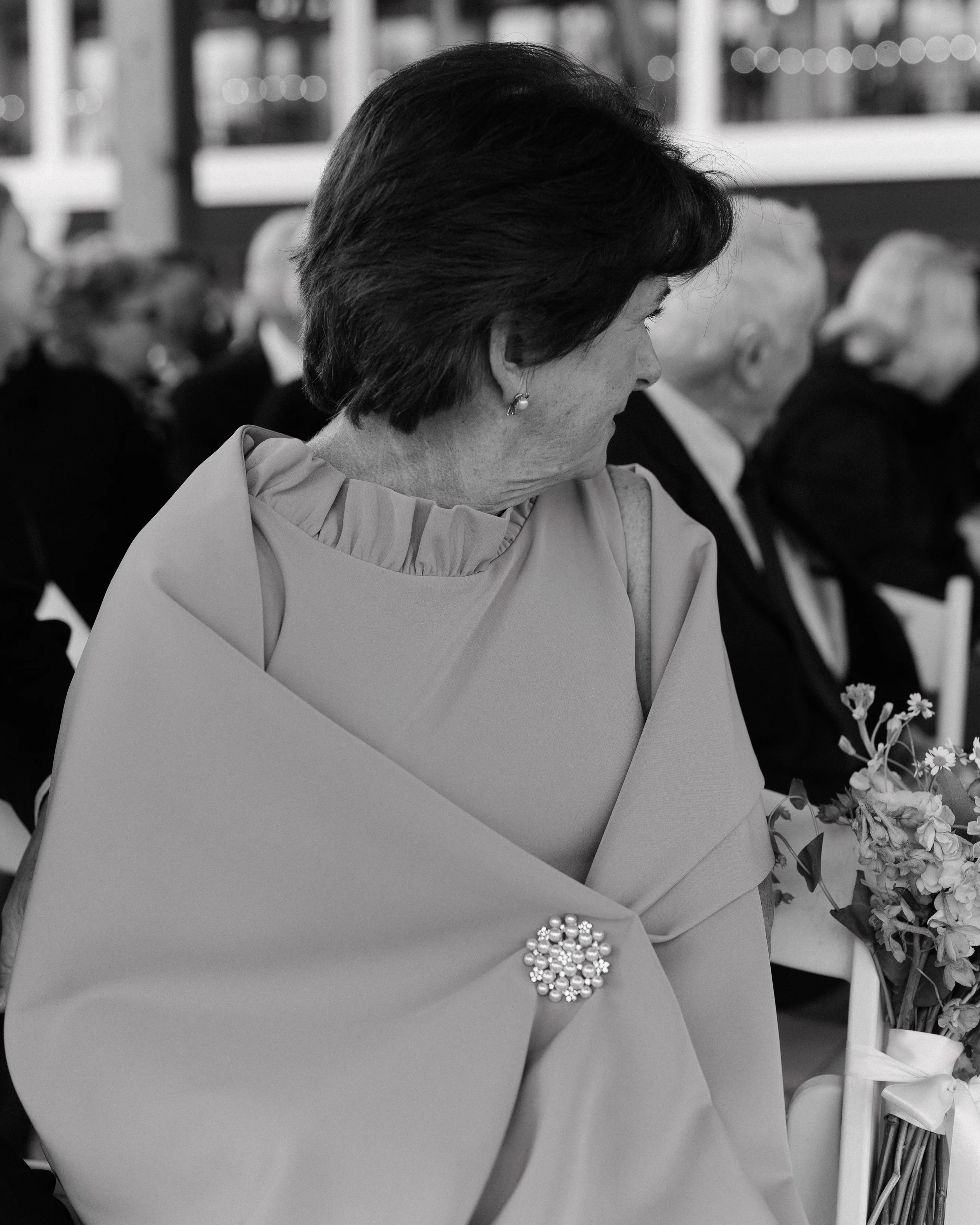 Black and white photograph of a woman wearing a formal dress with a large brooch, sitting in a row of people dressed formally, with some holding flowers, attending an event.