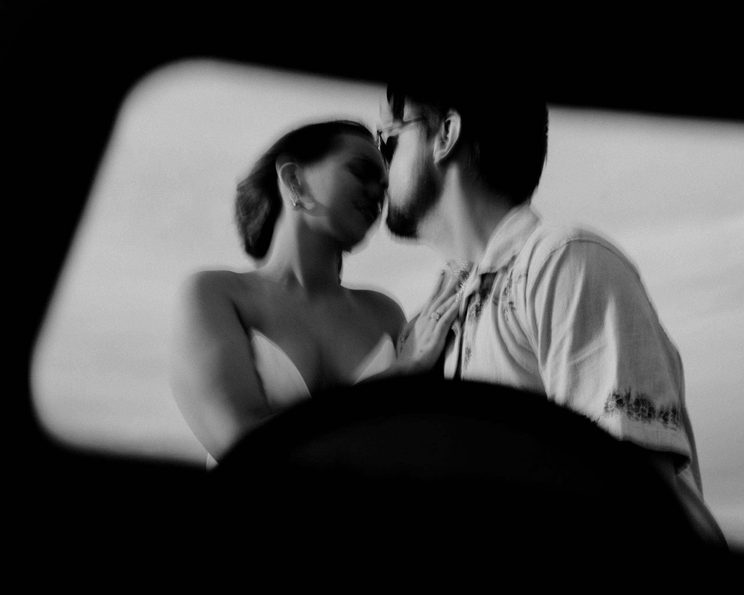 Black and white photo of a couple kissing, seen through a blurred frame. The woman is wearing a strapless dress, and the man is wearing a shirt with sunglasses.