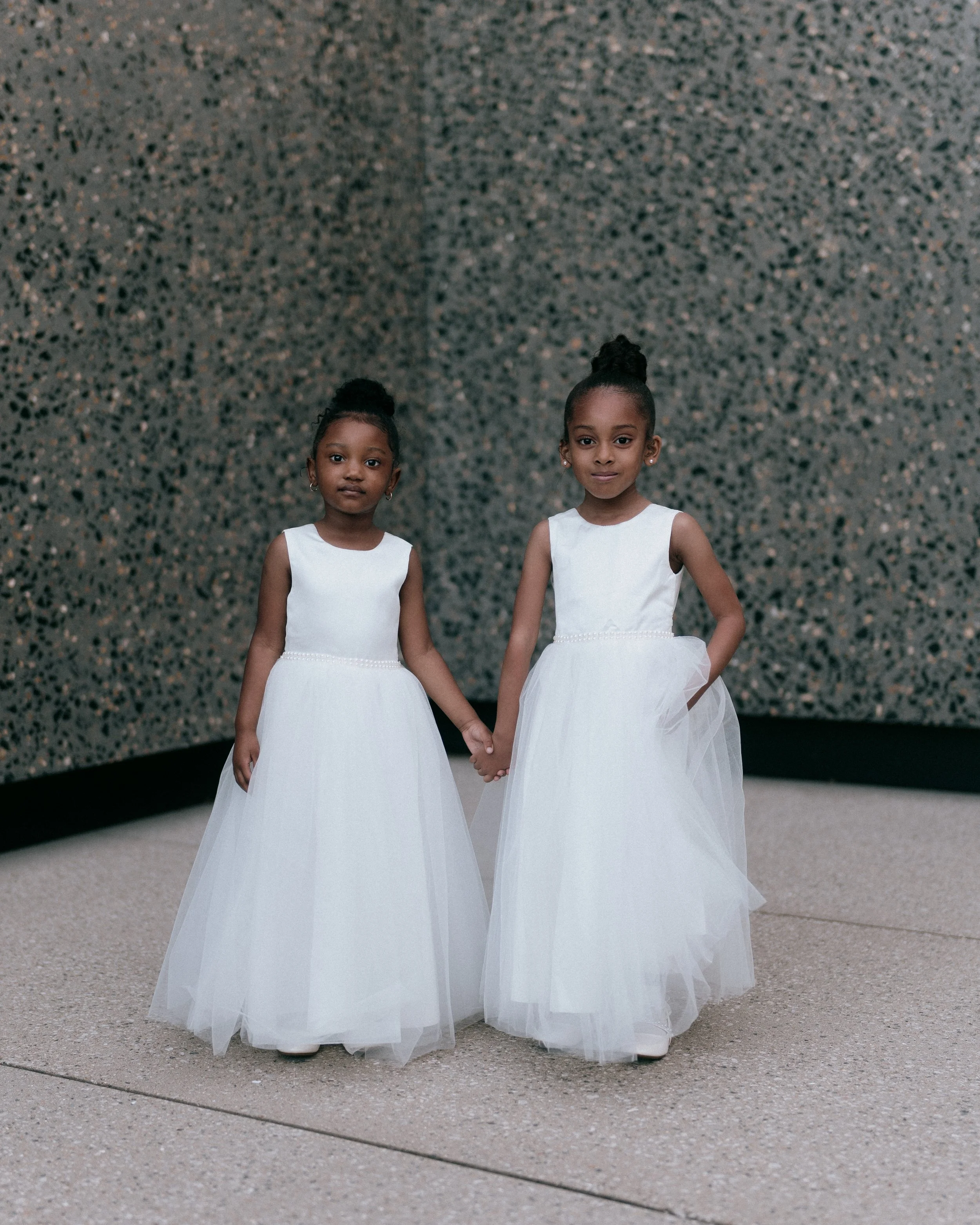 Two young girls in white dresses holding hands, standing against a textured wall.