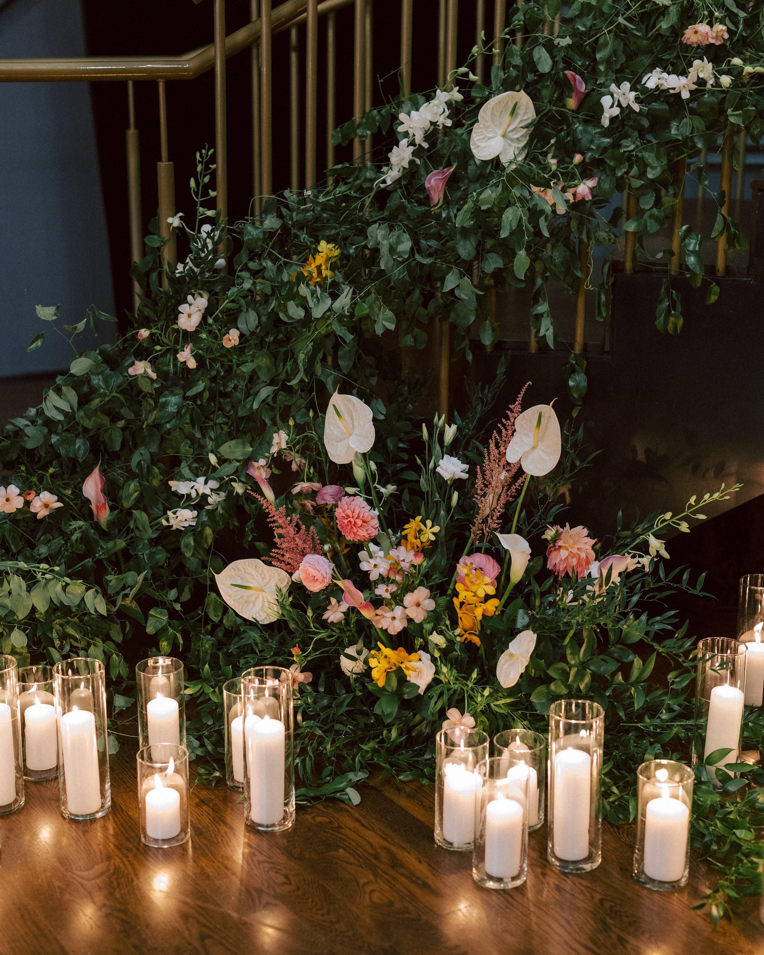 A floral arrangement with white calla lilies, pink and yellow flowers, and greenery, surrounded by white candles in glass holders on a wooden surface, set against a dark background.