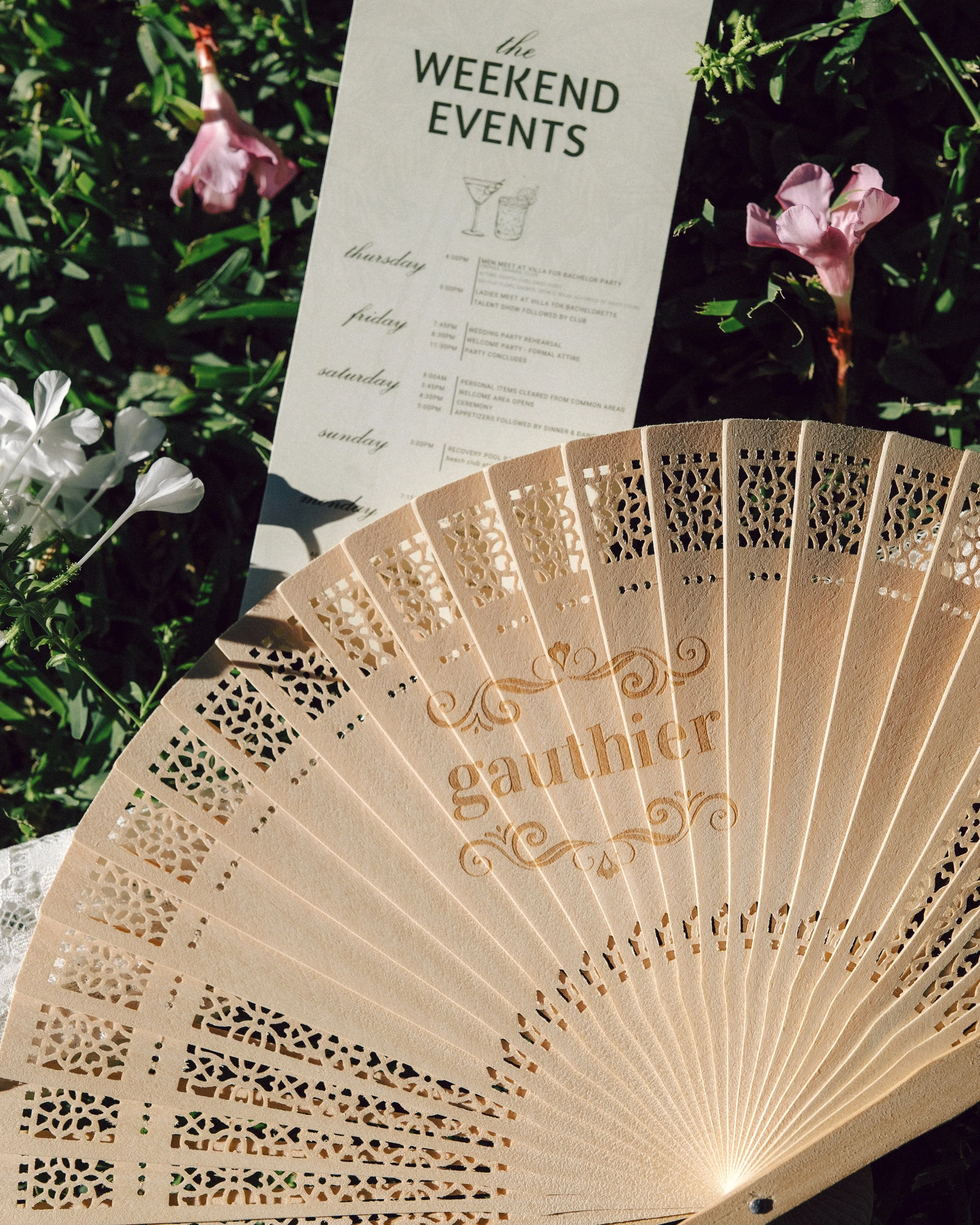 A decorative hand fan with the word 'Gaulther' on it, placed on grass beside a printed events schedule titled 'Weekend Events' for Thursday through Sunday, surrounded by pink and white flowers.