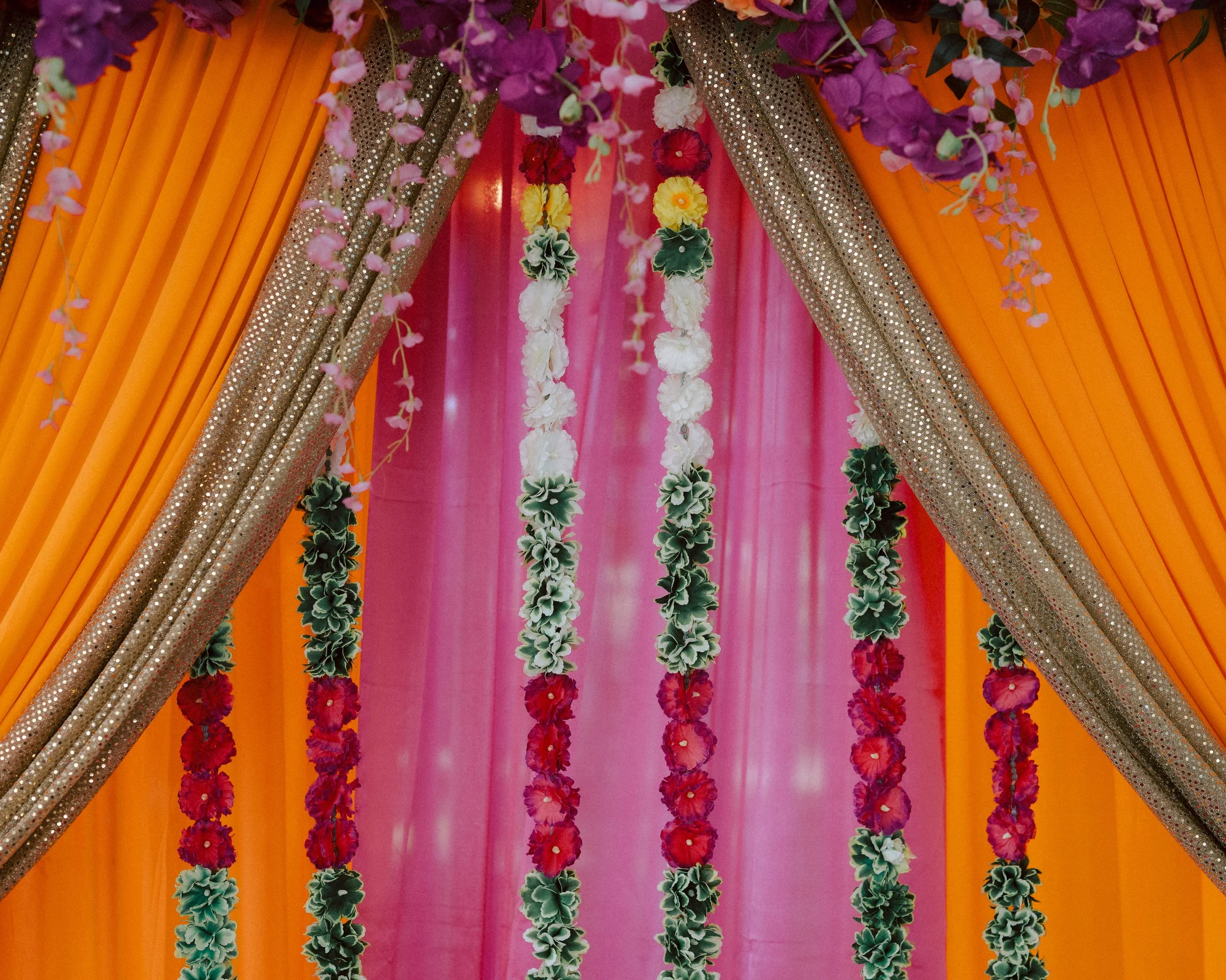 Decorative floral drapes with orange, gold, pink, green, and purple colors, accented with hanging flower garlands.