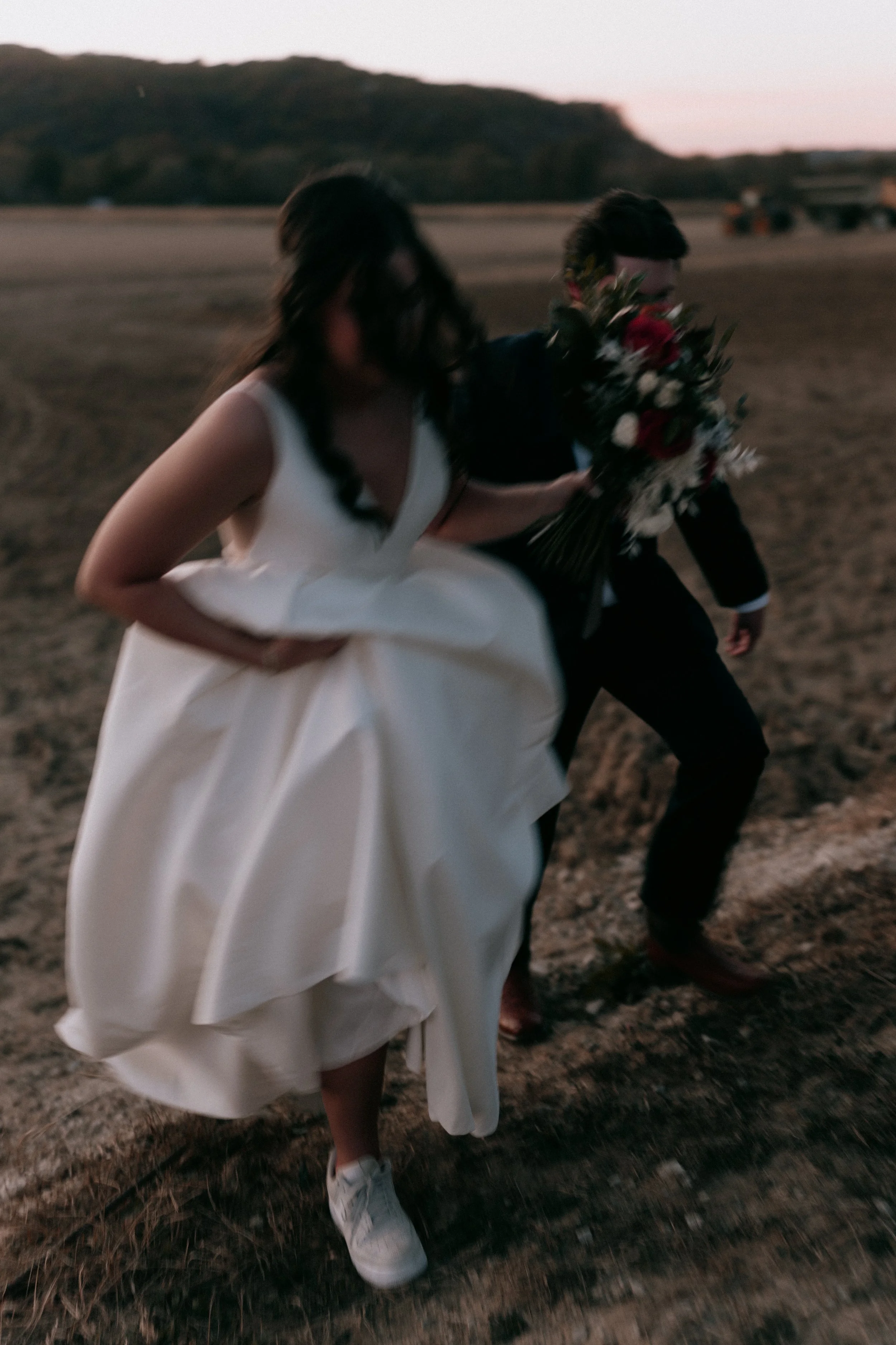 Blurry photo of a bride and groom walking in a field, bride wearing sneakers and holding bouquet, during sunset.