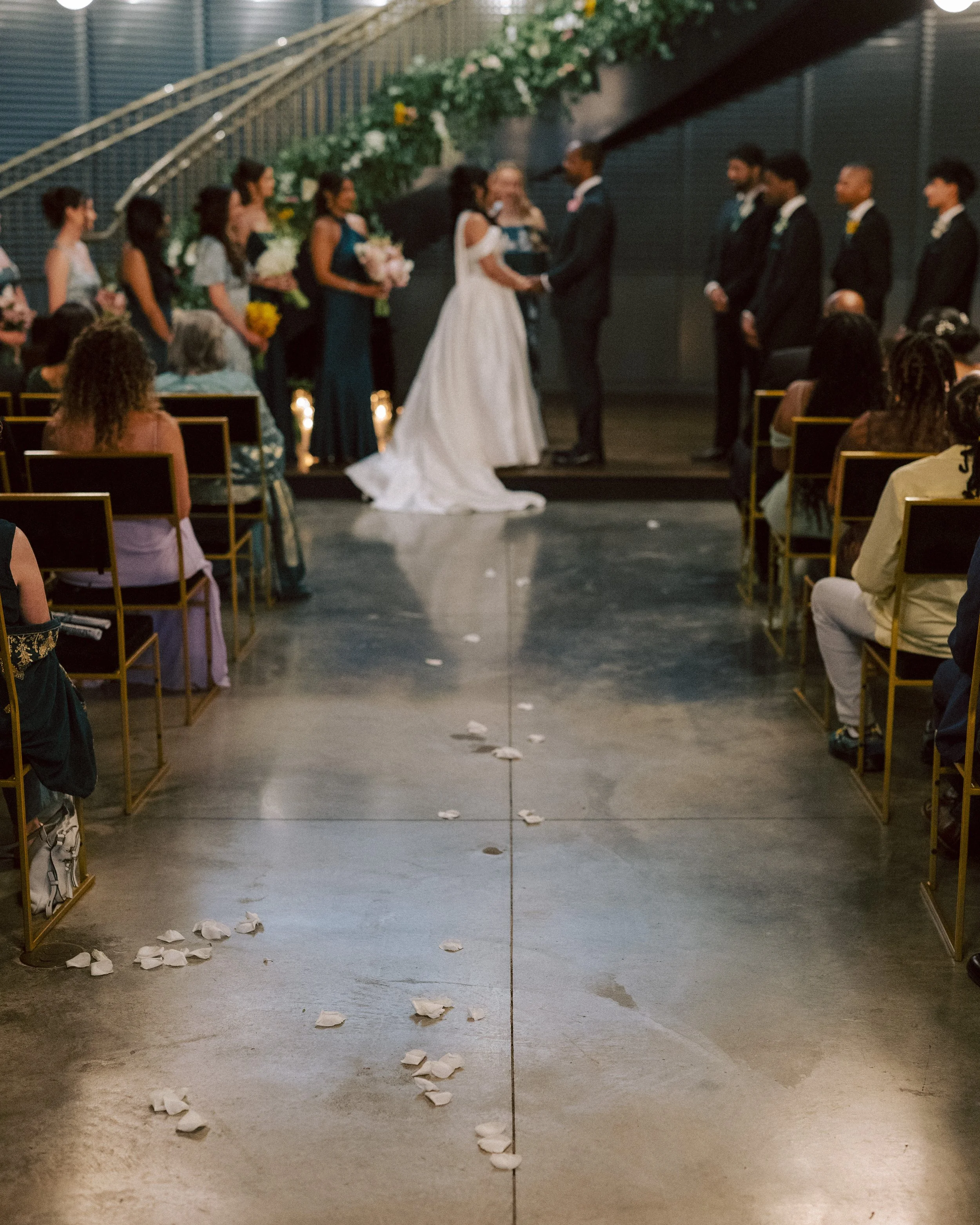 Couple getting married at an indoor wedding ceremony with guests seated on both sides, flower petals on the floor, and wedding party standing nearby.