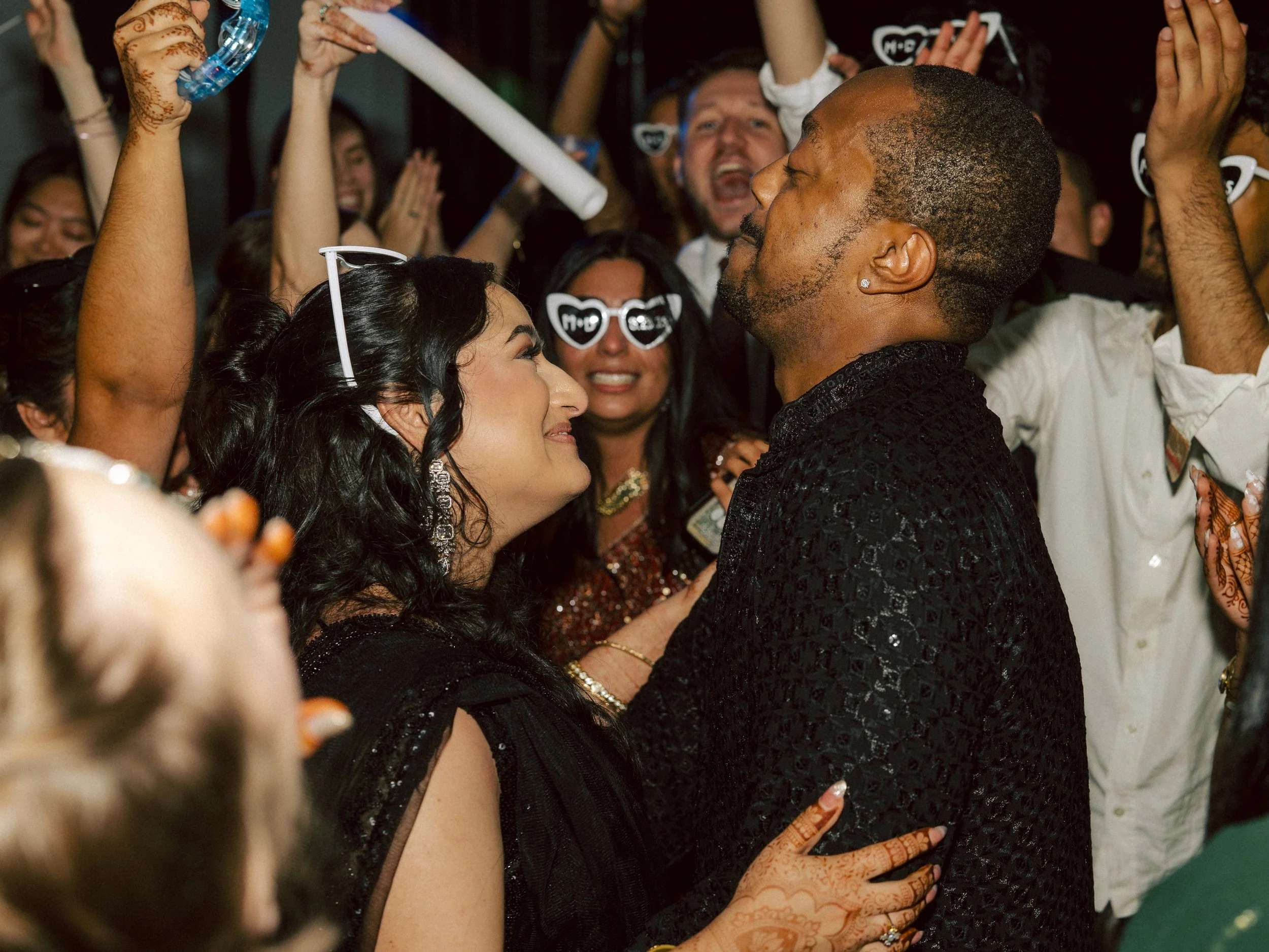 A group of people celebrating at a party, with two individuals in the foreground feeling emotional, surrounded by friends wearing party accessories and glasses, and holding drinks.