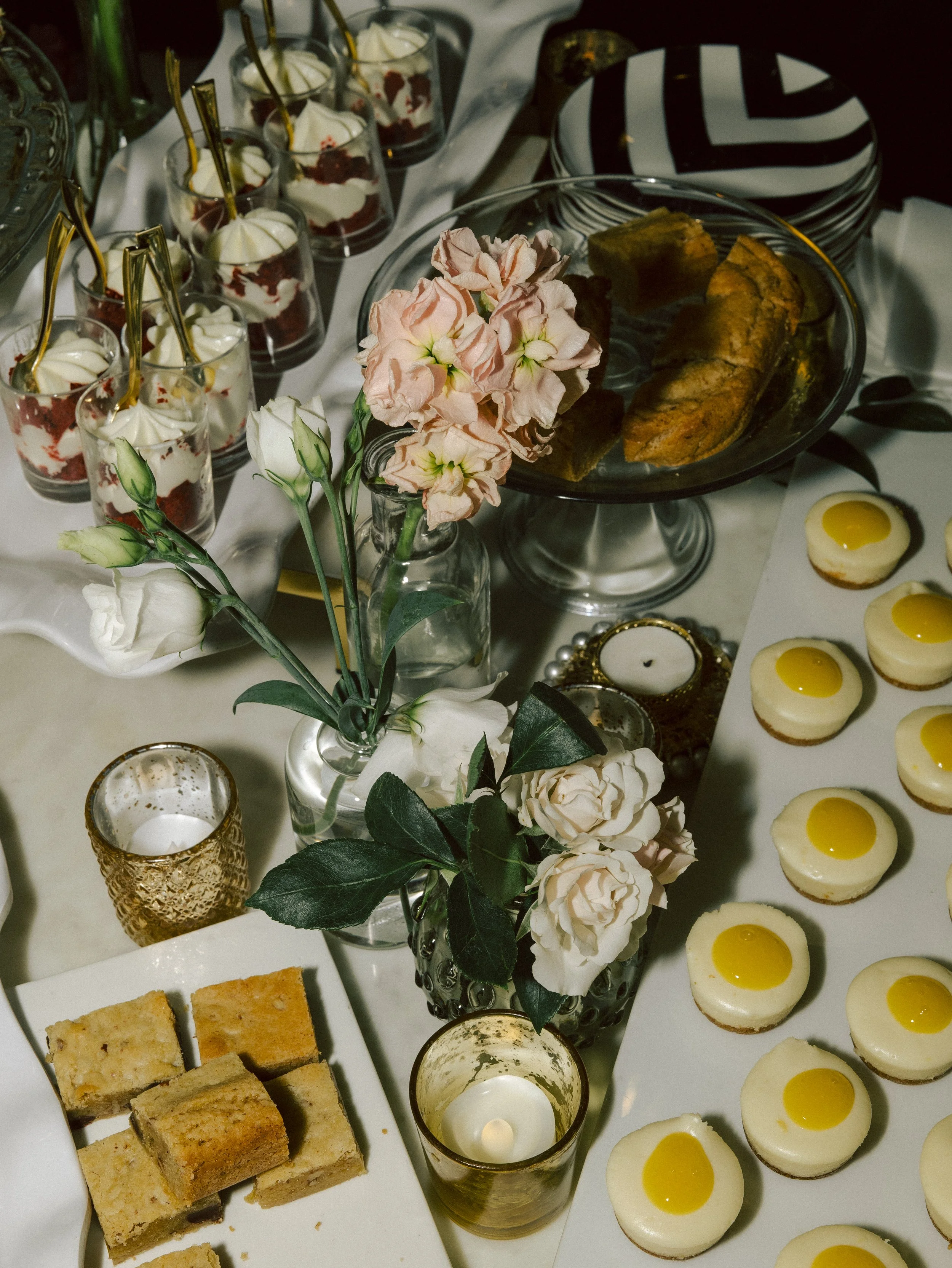 Table with desserts, flowers, candles, and plates, including small glasses with whipped cream and strawberries, cookies, and mini cheesecakes topped with yellow glaze.