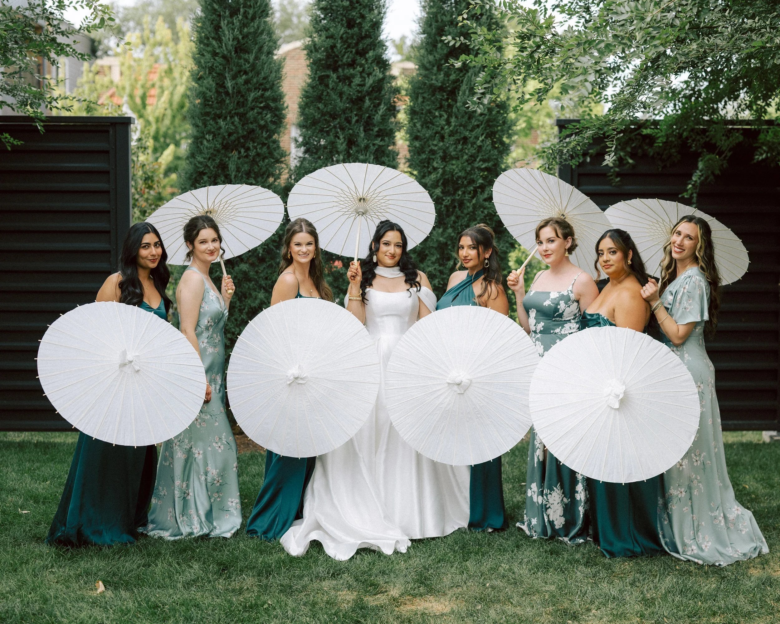 A bride and seven bridesmaids standing on grass, holding white paper parasols, outdoors with green trees and a black fence in the background.